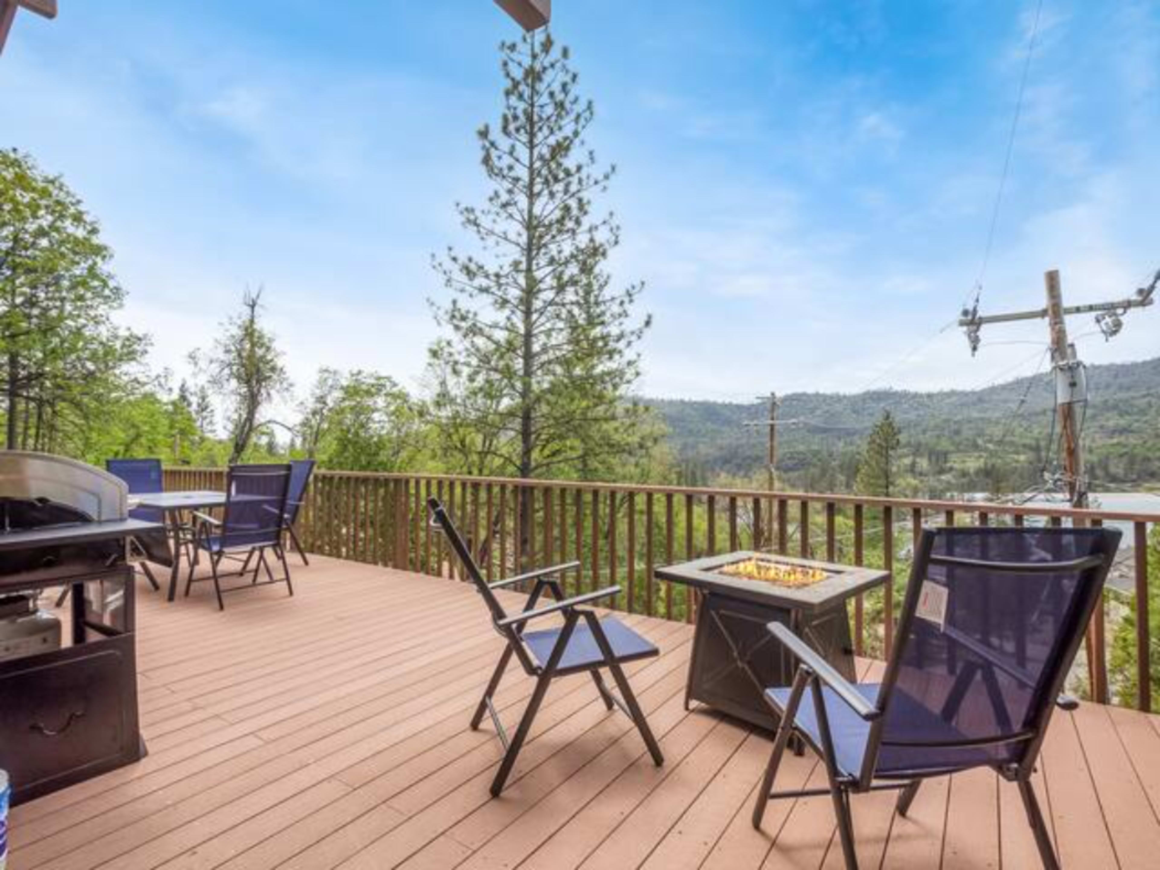 A wooden deck with black patio chairs and a fire pit, overlooking a forested landscape and distant hills.