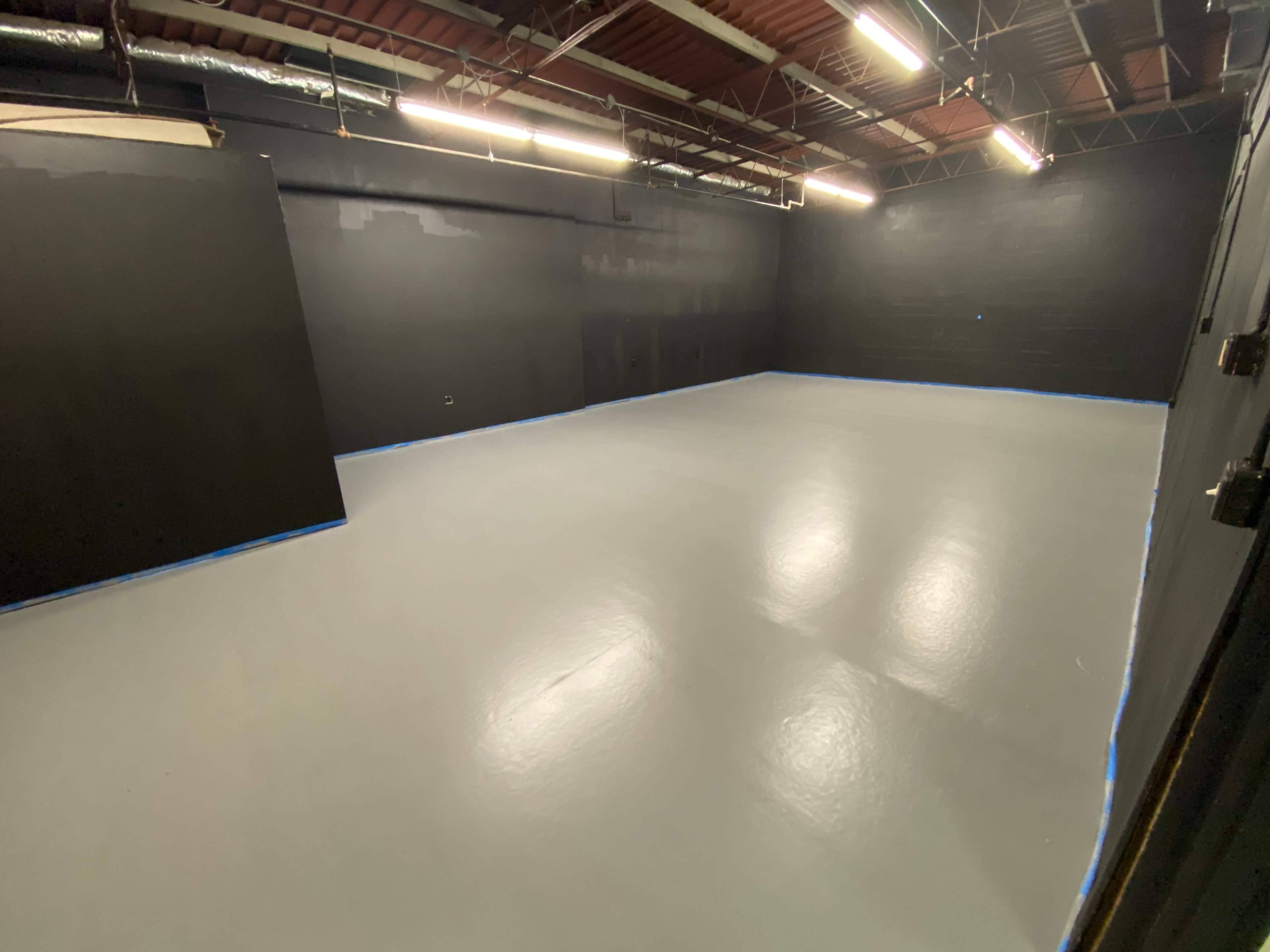 A large, empty room with a gray floor and black walls, illuminated by overhead lights.