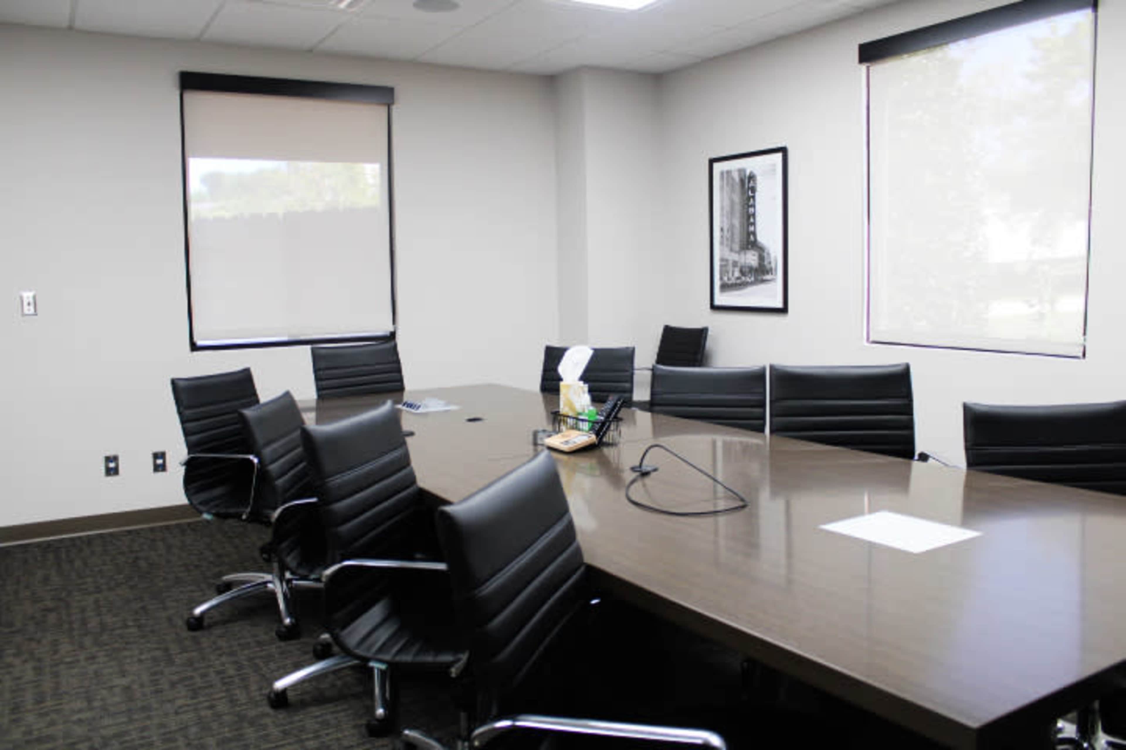 A conference room features a long table surrounded by black chairs and has windows with shades partially closed.