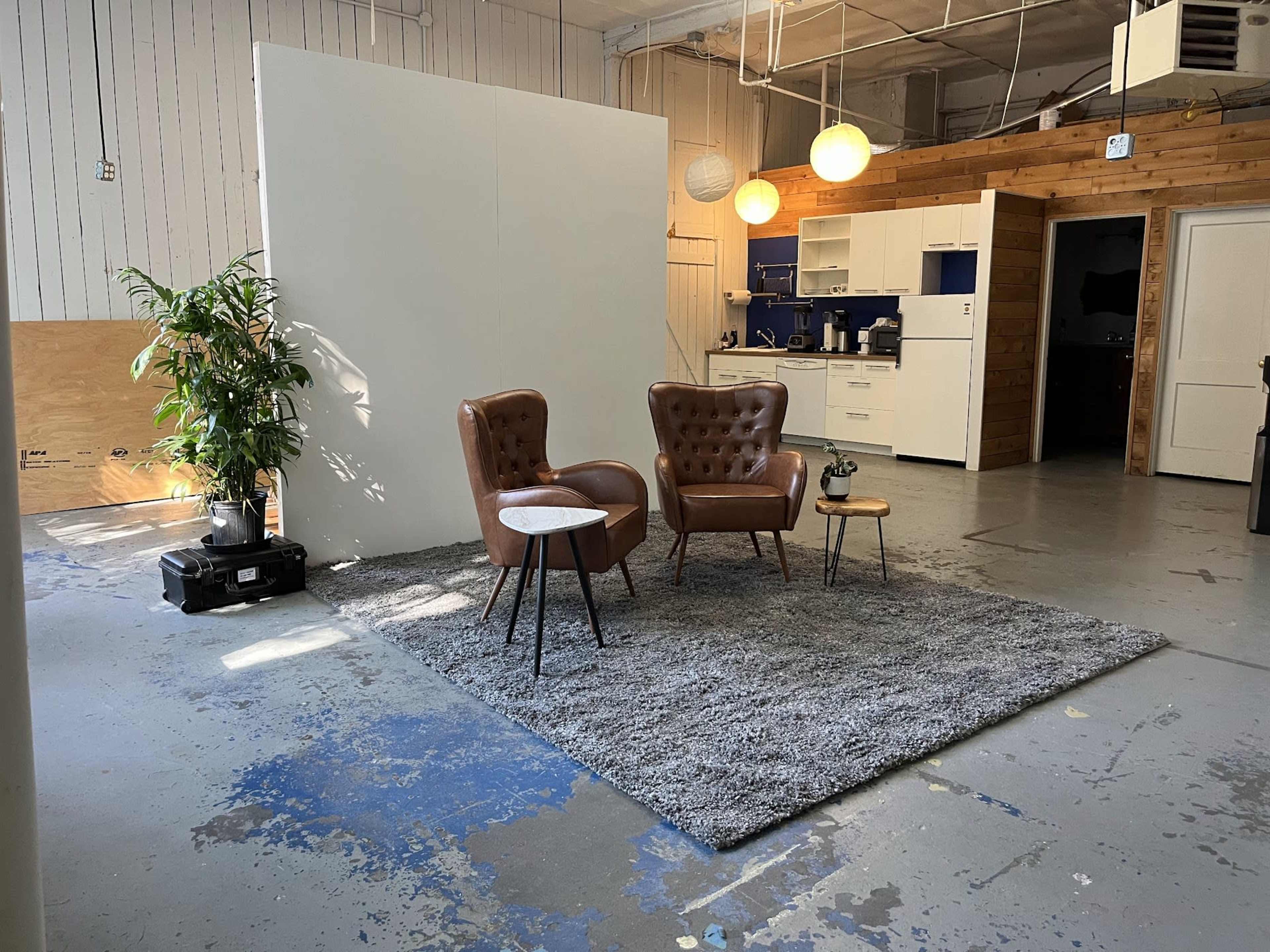 A sparse interior space featuring two brown leather chairs, a small side table, and a large rug, with a white wall in the background and a kitchen area visible to one side.