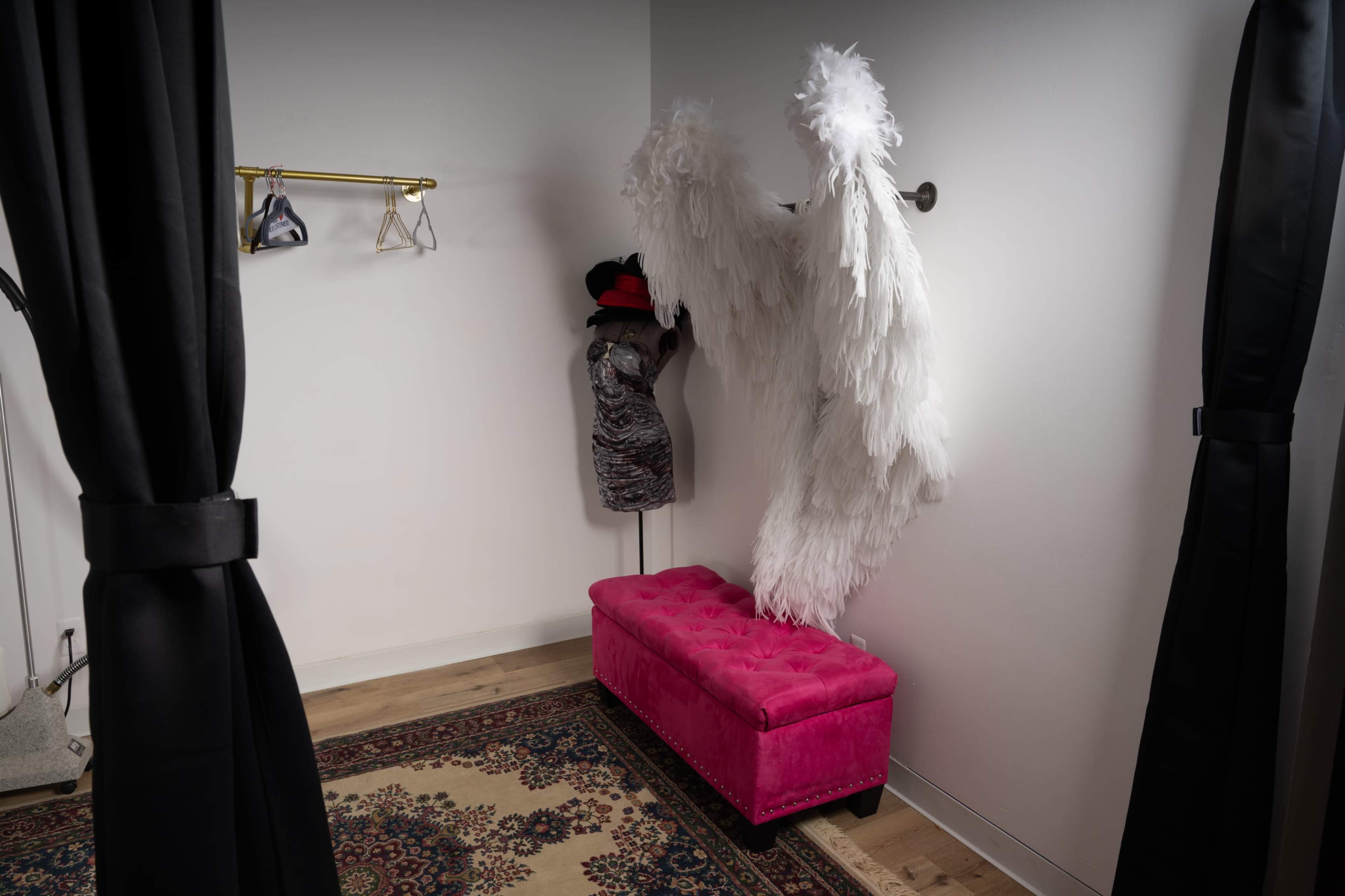 A small dressing room features a pink tufted bench, a mannequin displaying a dress, and large white feather wings hanging on the wall.
