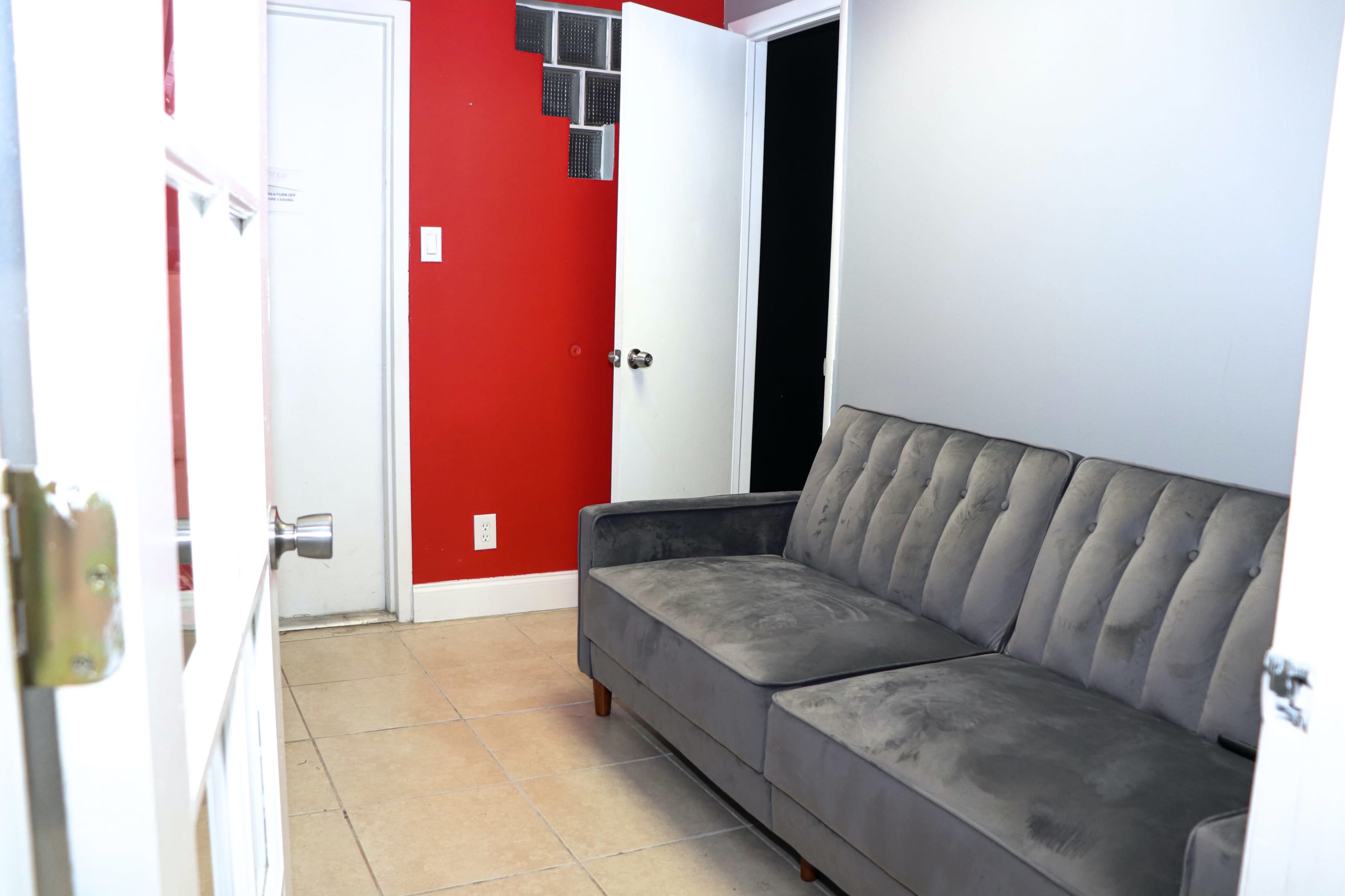 A gray tufted sofa is positioned against a wall in a small room with red and white walls, featuring two doors and a tiled floor.