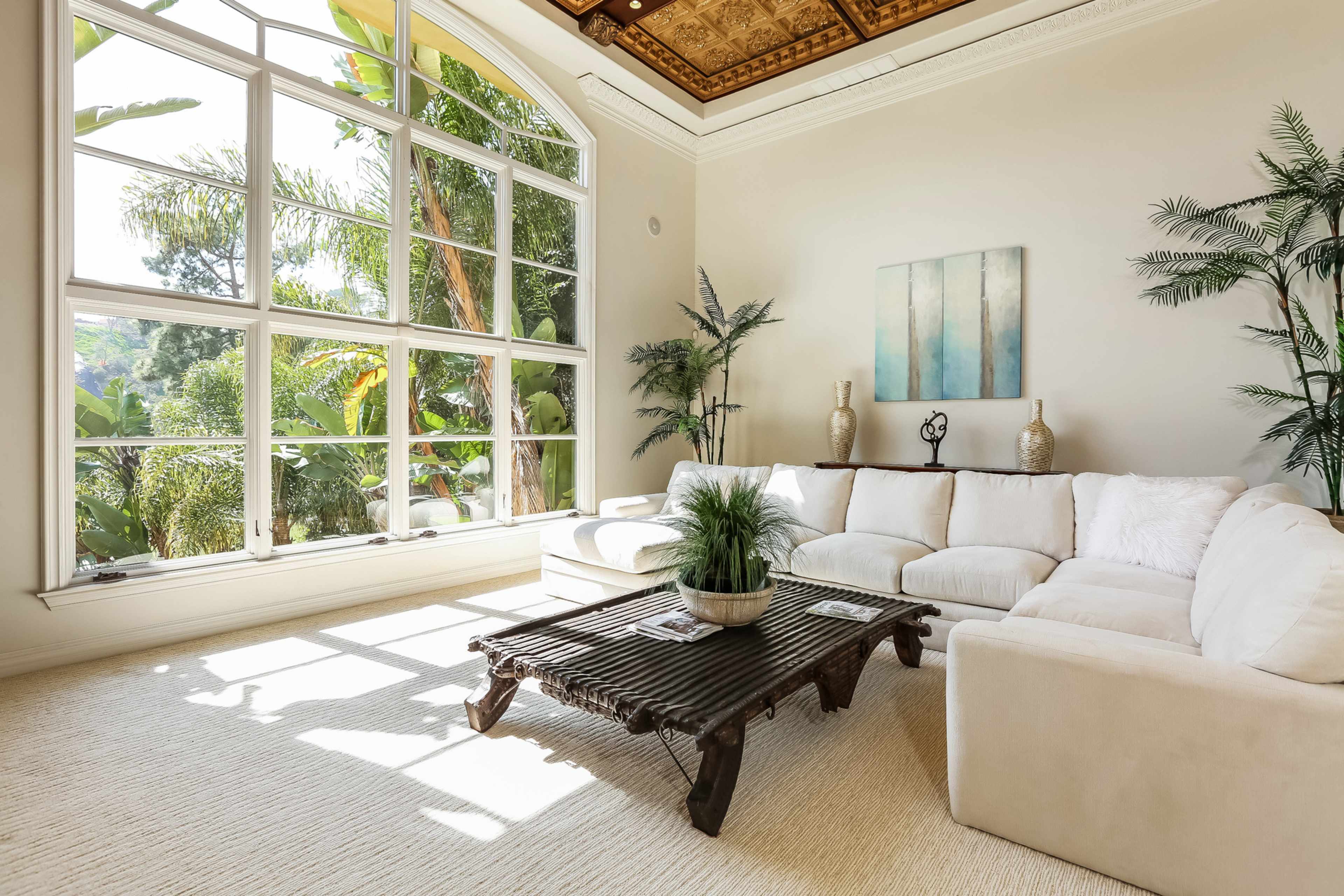A bright living room features a large window with greenery outside, a white sectional sofa, and a dark wooden coffee table.