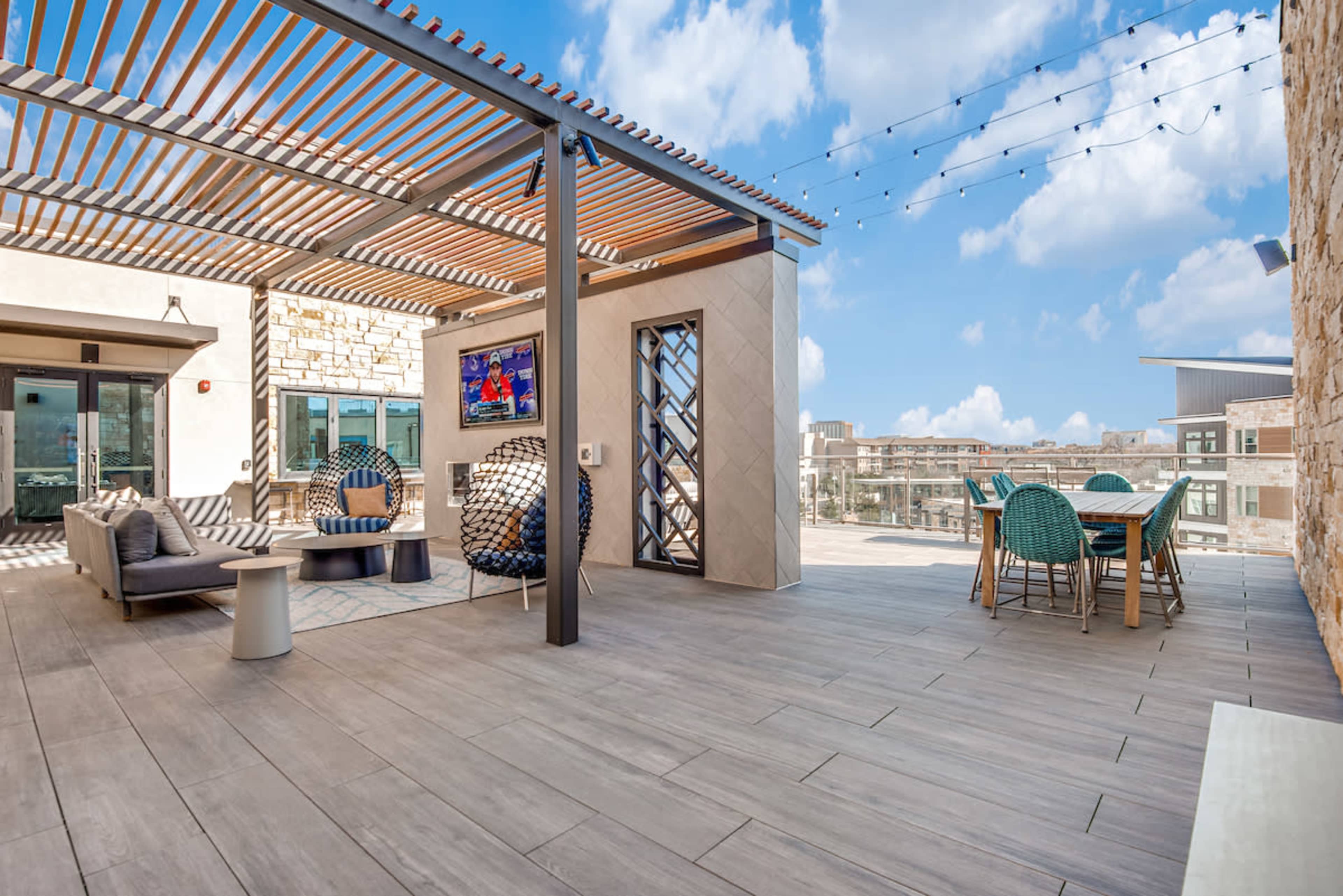 The rooftop terrace features modern outdoor furniture, a television, and string lights against a backdrop of blue skies and clouds.