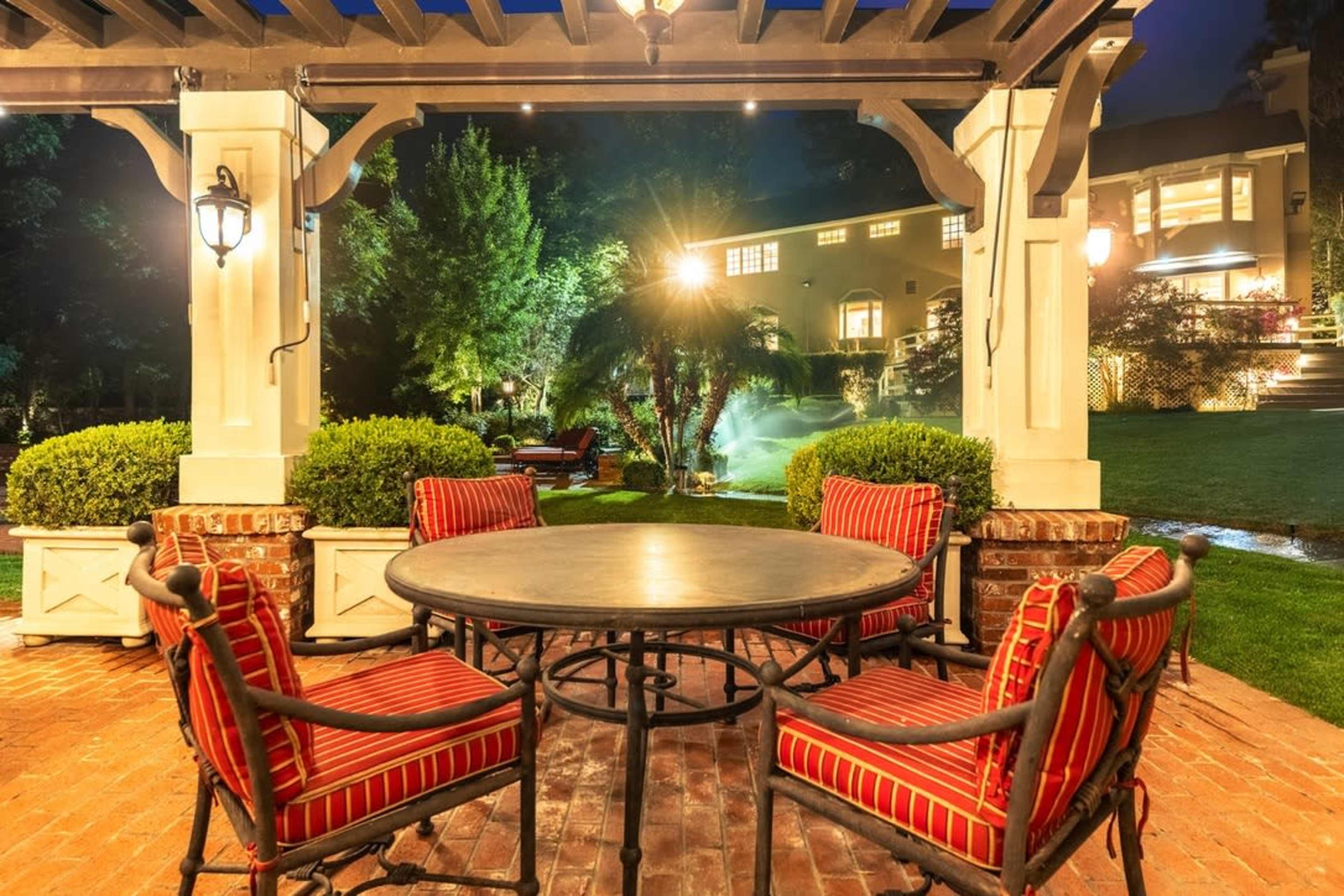 A patio area features a round table surrounded by four striped chairs, with lush greenery and a well-lit house in the background.