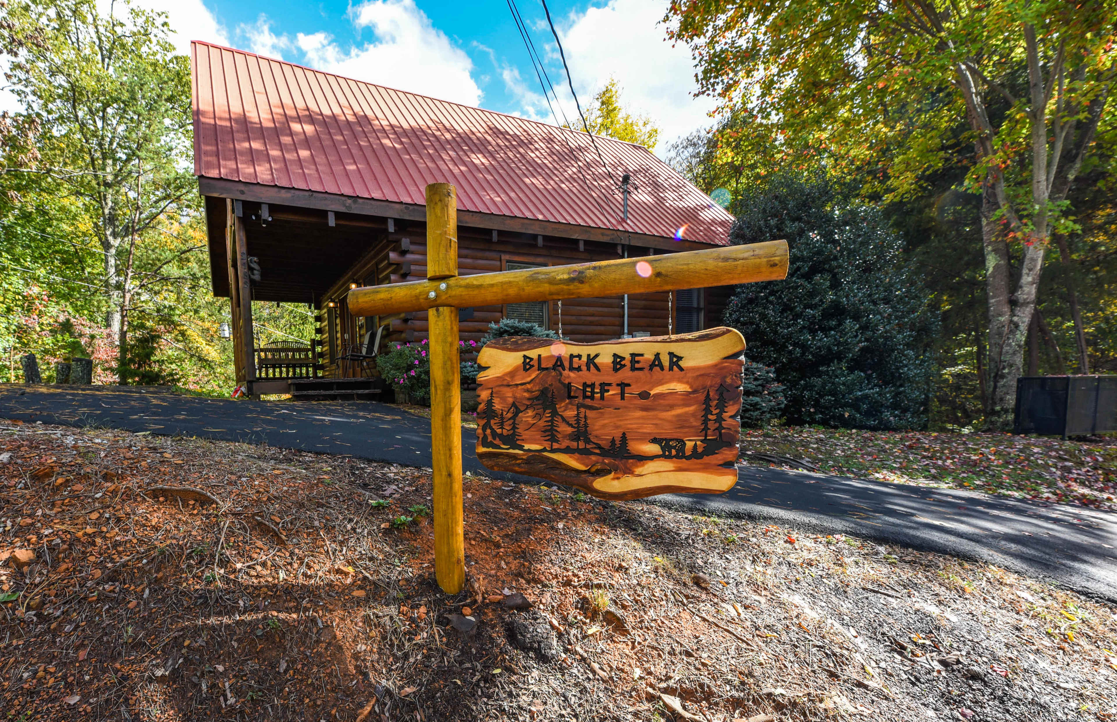 A wooden sign reading "Black Bear Left" stands at the entrance to a log cabin with a red metal roof surrounded by trees.