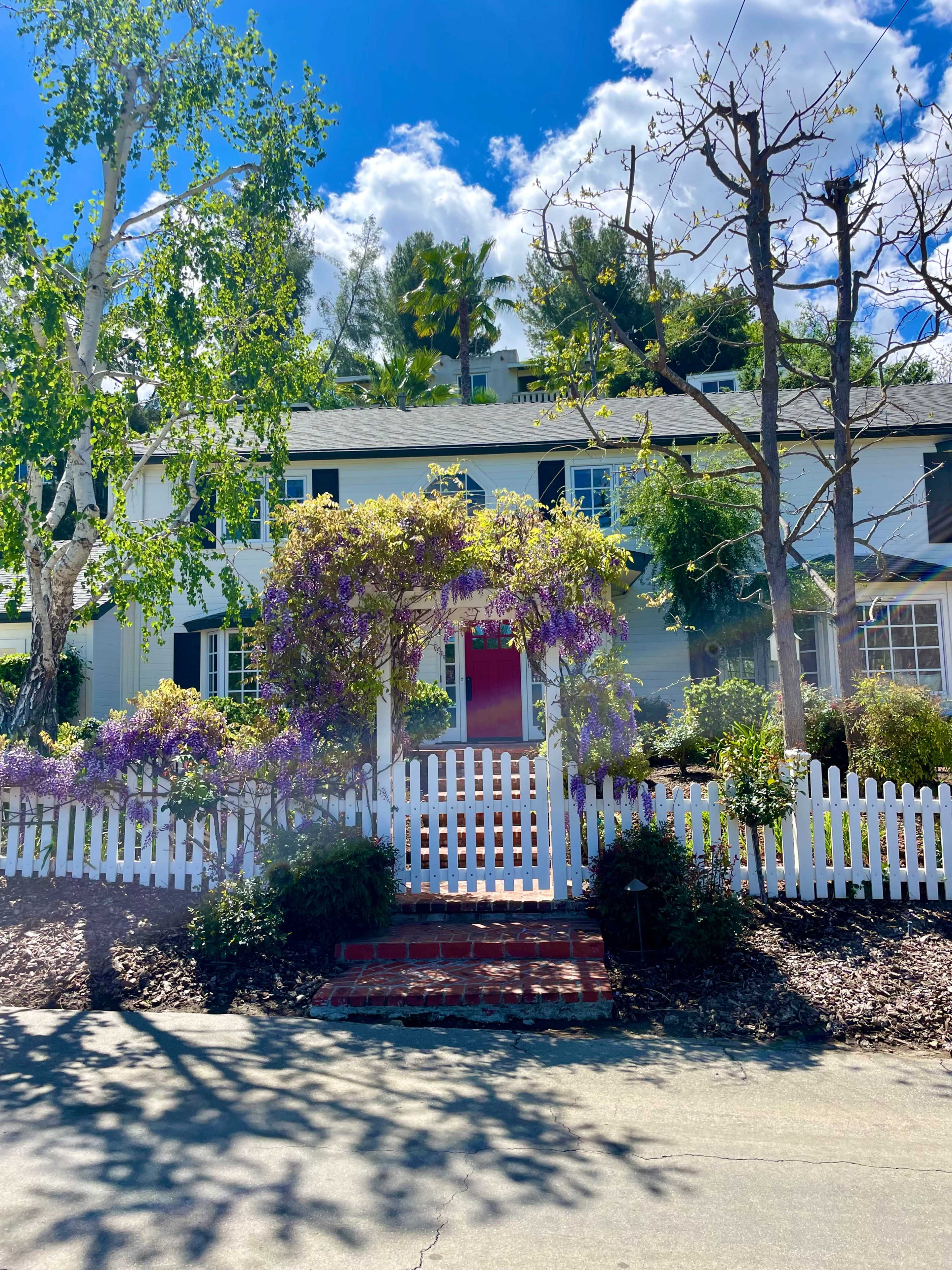 A two-story white house with a red door is surrounded by lush greenery and a white picket fence, with purple flowering vines and a paved walkway leading to the entrance.