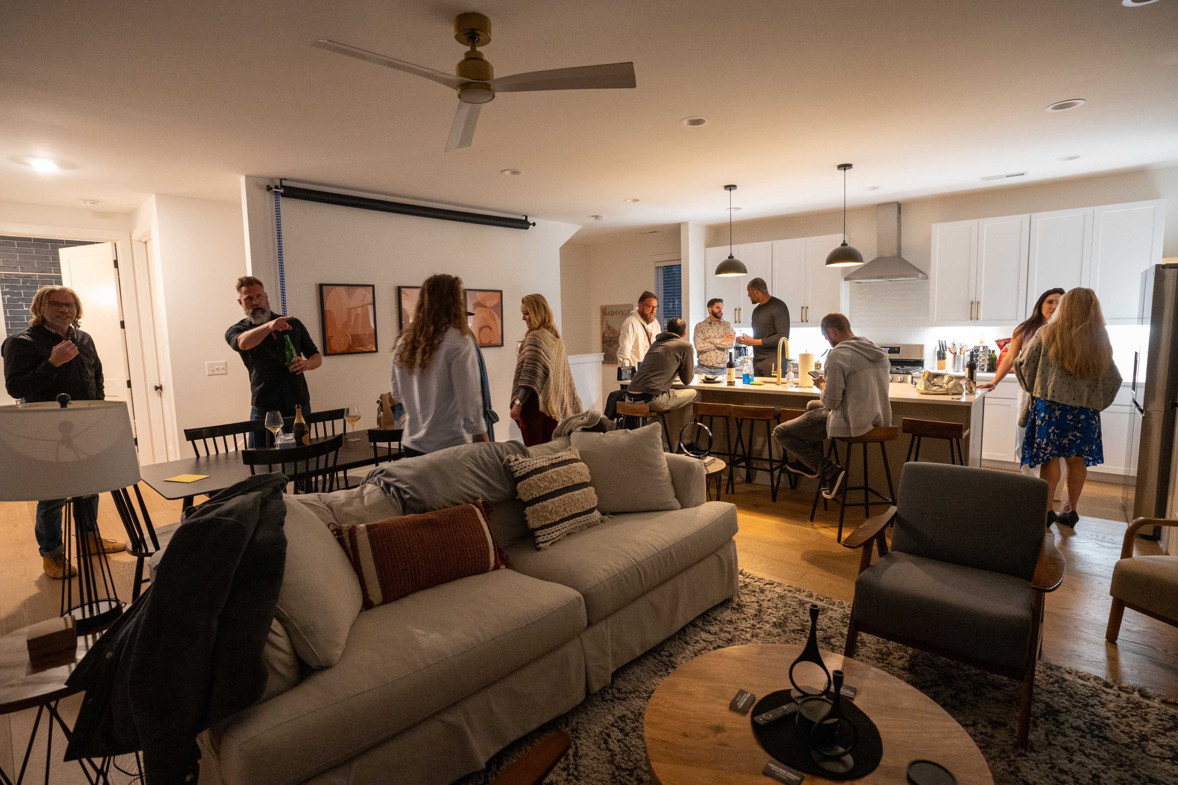 A group of people are gathered in a modern, well-lit living space, with some mingling near a kitchen island while others sit on furniture.