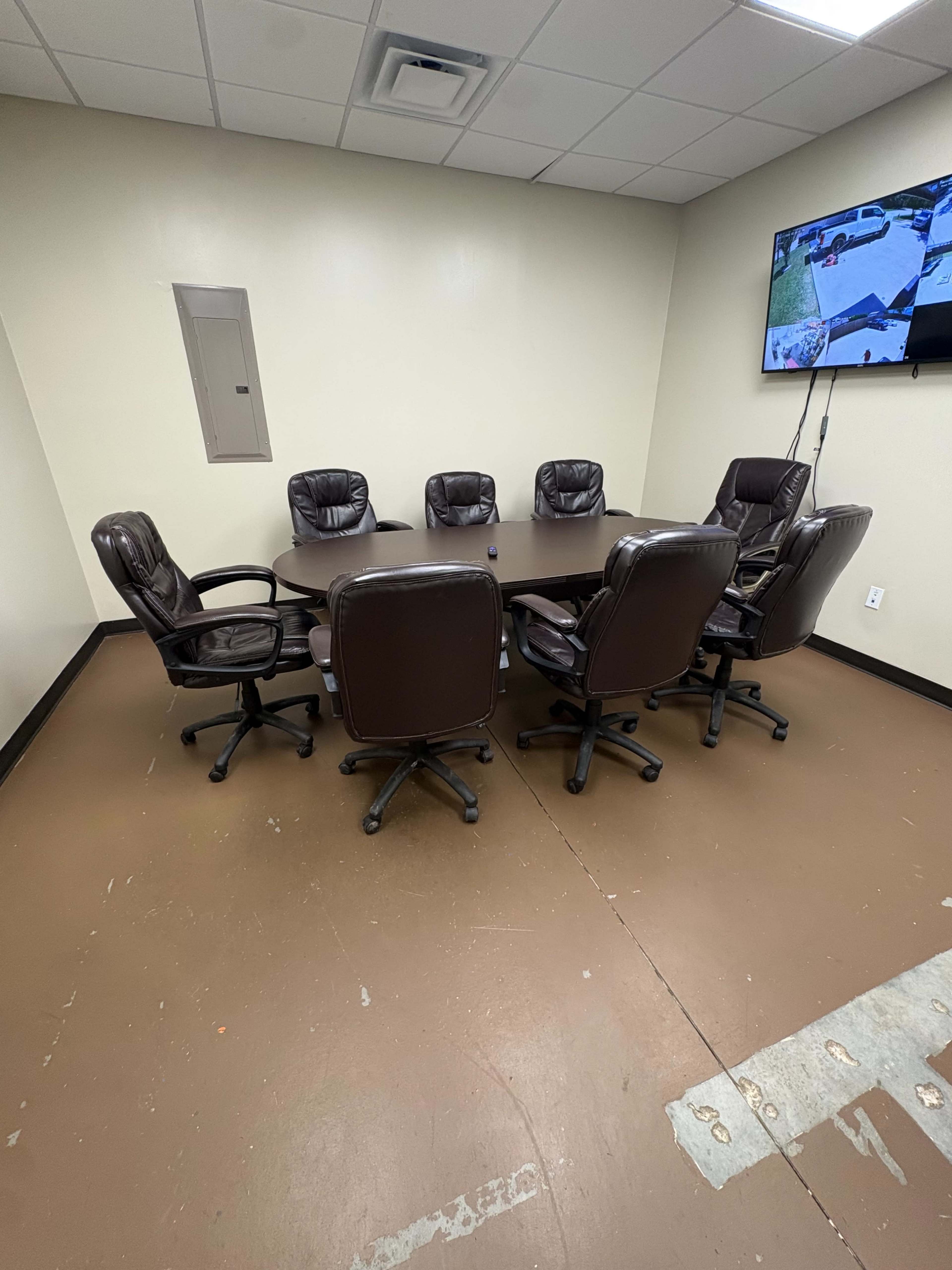A round conference table surrounded by eight padded office chairs is positioned in a plain meeting room with a television mounted on the wall.