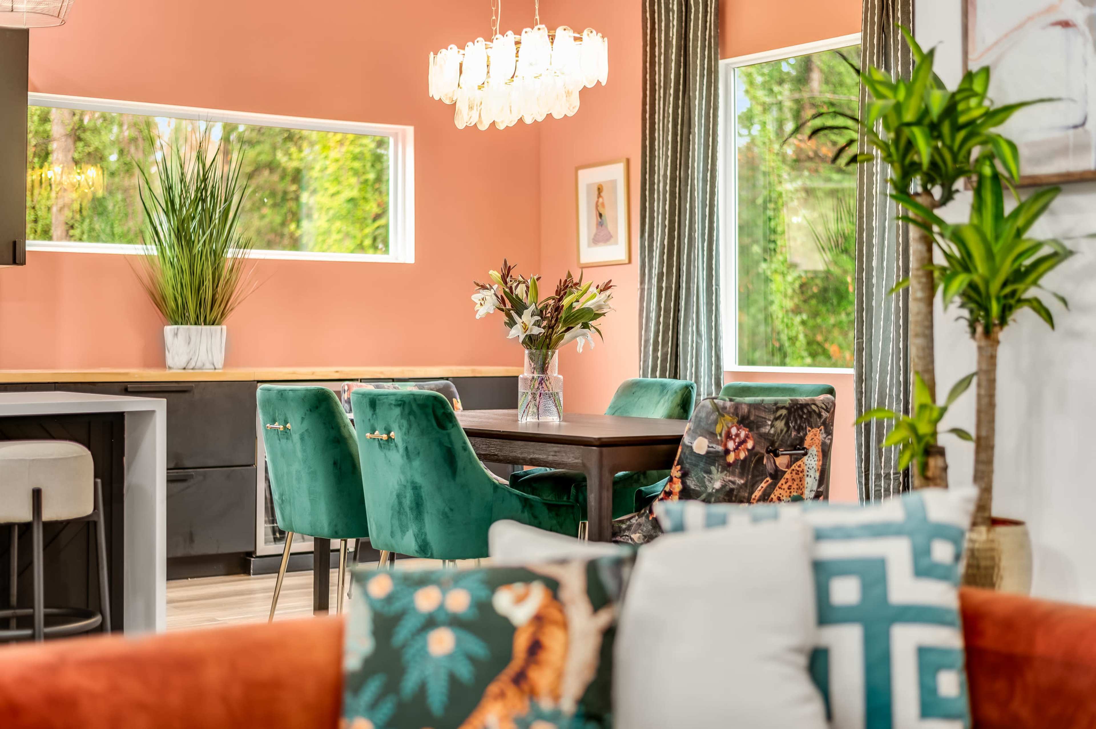 A modern dining area features a dark wooden table surrounded by green velvet chairs, with a floral centerpiece and large windows letting in natural light against a peach-painted wall.