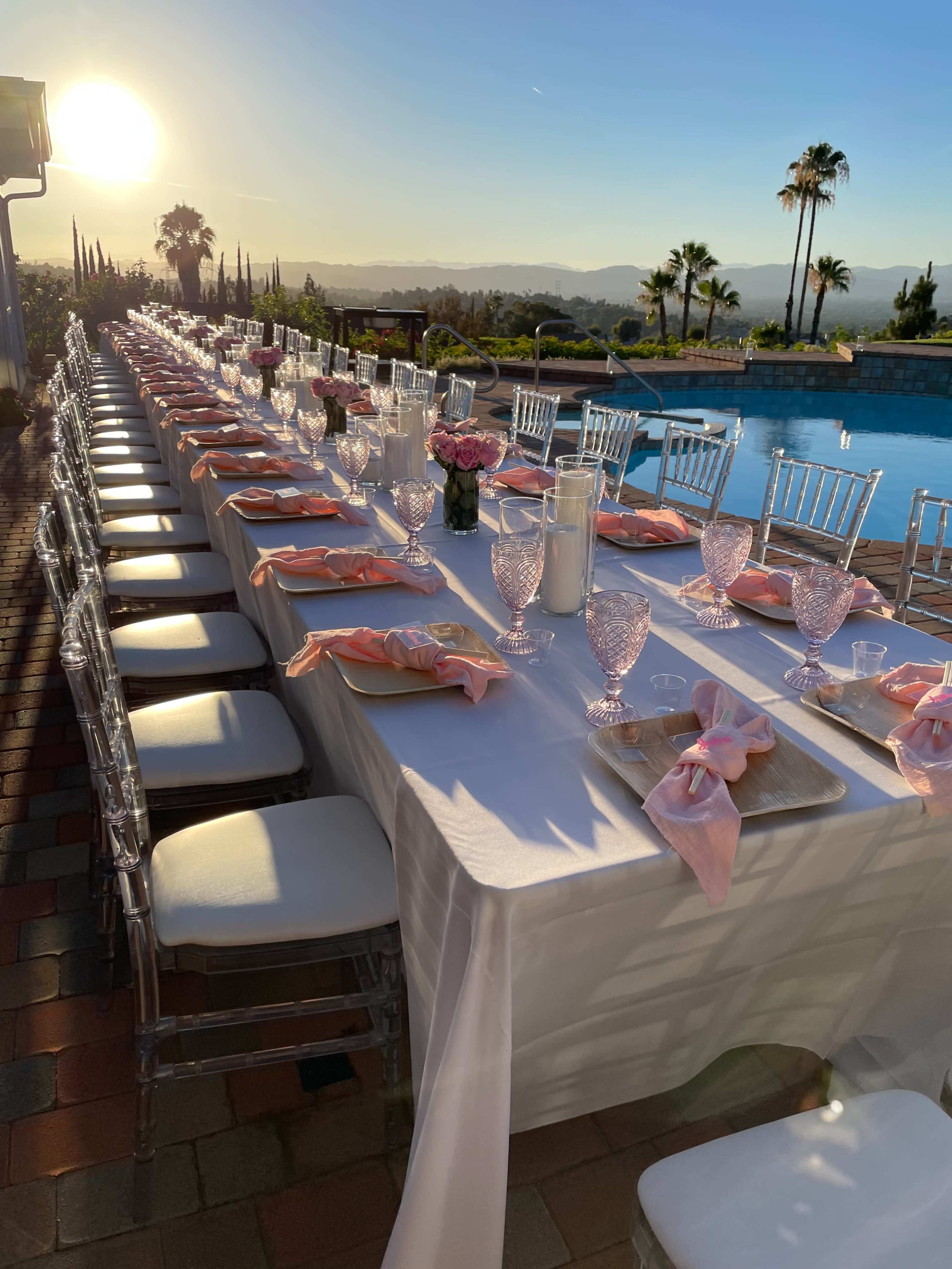 A long banquet table is set with pink napkins and floral centerpieces, overlooking a pool and a sunset in the background.