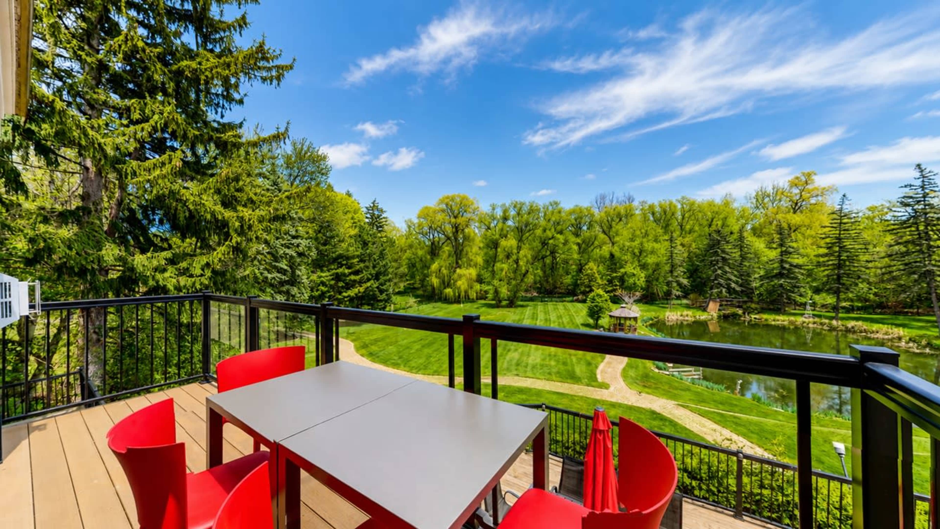 A deck with a table and red chairs overlooks a lush green landscape and a pond surrounded by trees.