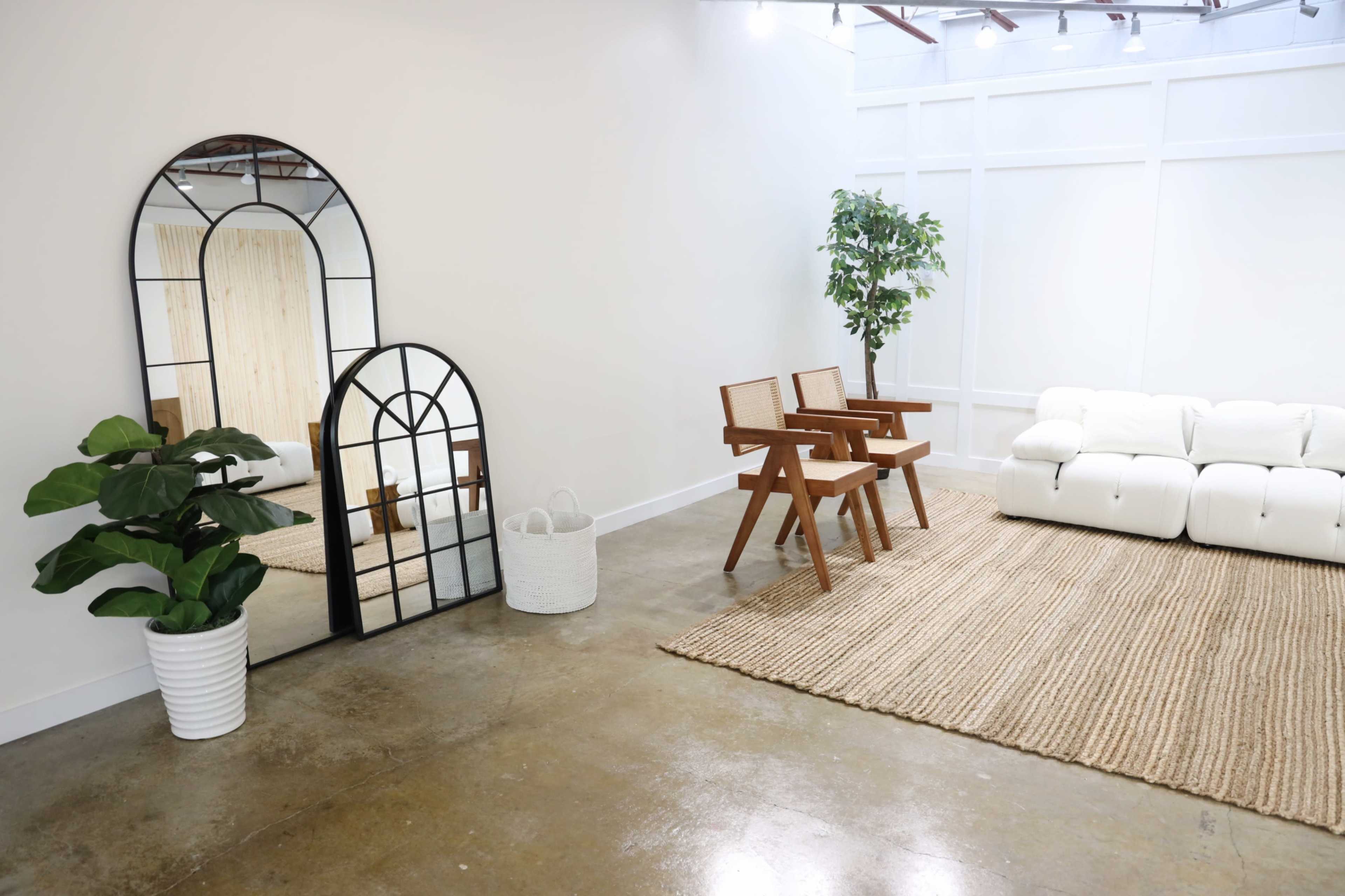 A well-lit room features a white sofa, two wooden chairs, a large mirror, a potted plant, and a woven rug on a polished concrete floor.