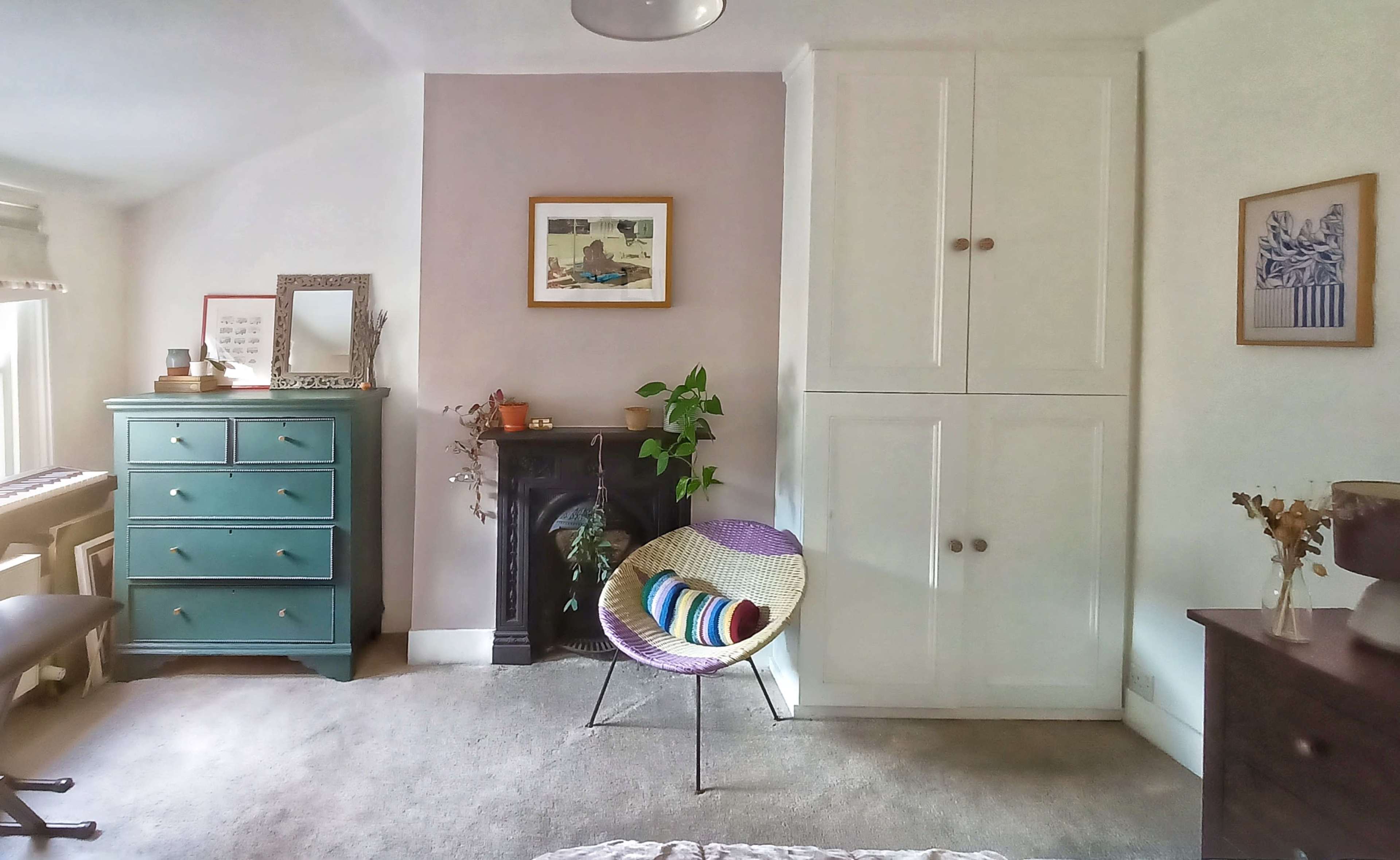 A bedroom featuring a light color scheme, a teal dresser, a wall-mounted artwork, a fireplace, and a woven chair.