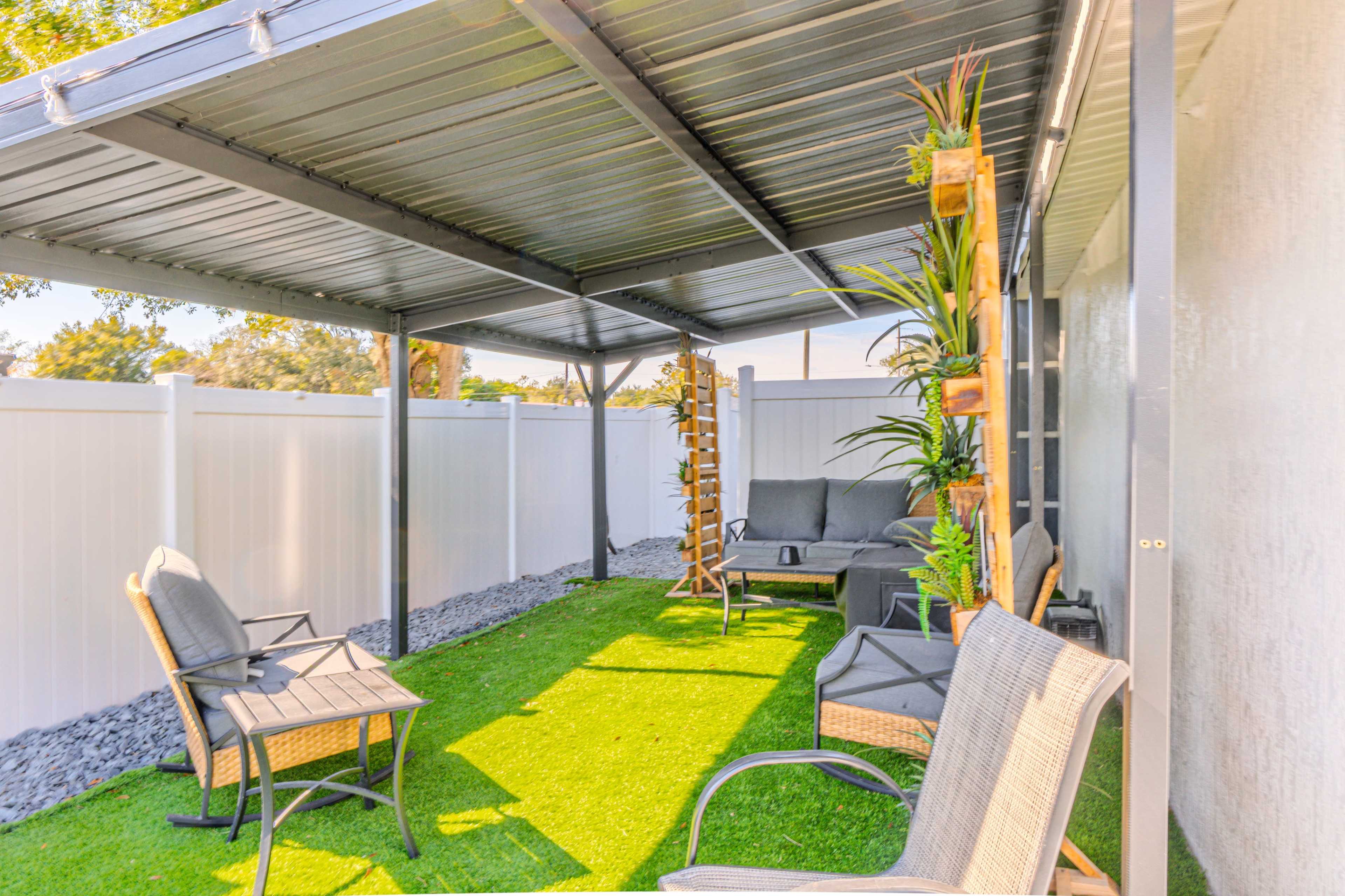The image shows a covered outdoor seating area with black chairs, a small table, and decorative plants, surrounded by white fences and artificial grass.