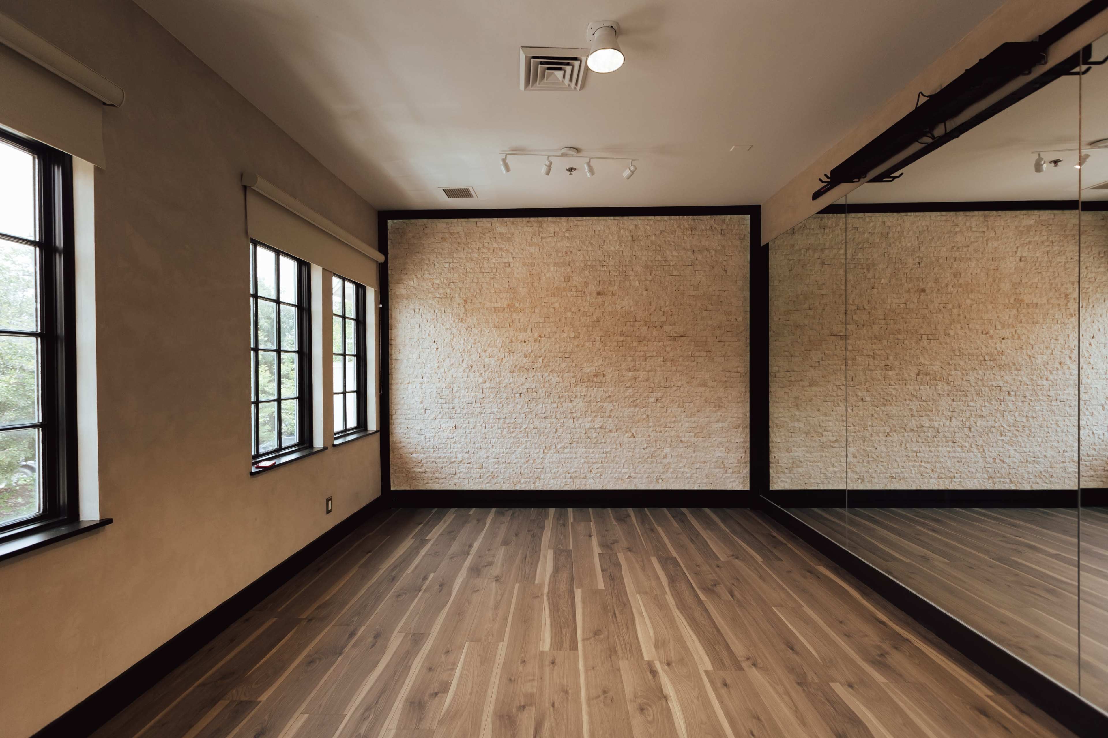 A spacious room with hardwood flooring, a textured stone wall, large windows, and one mirrored wall.