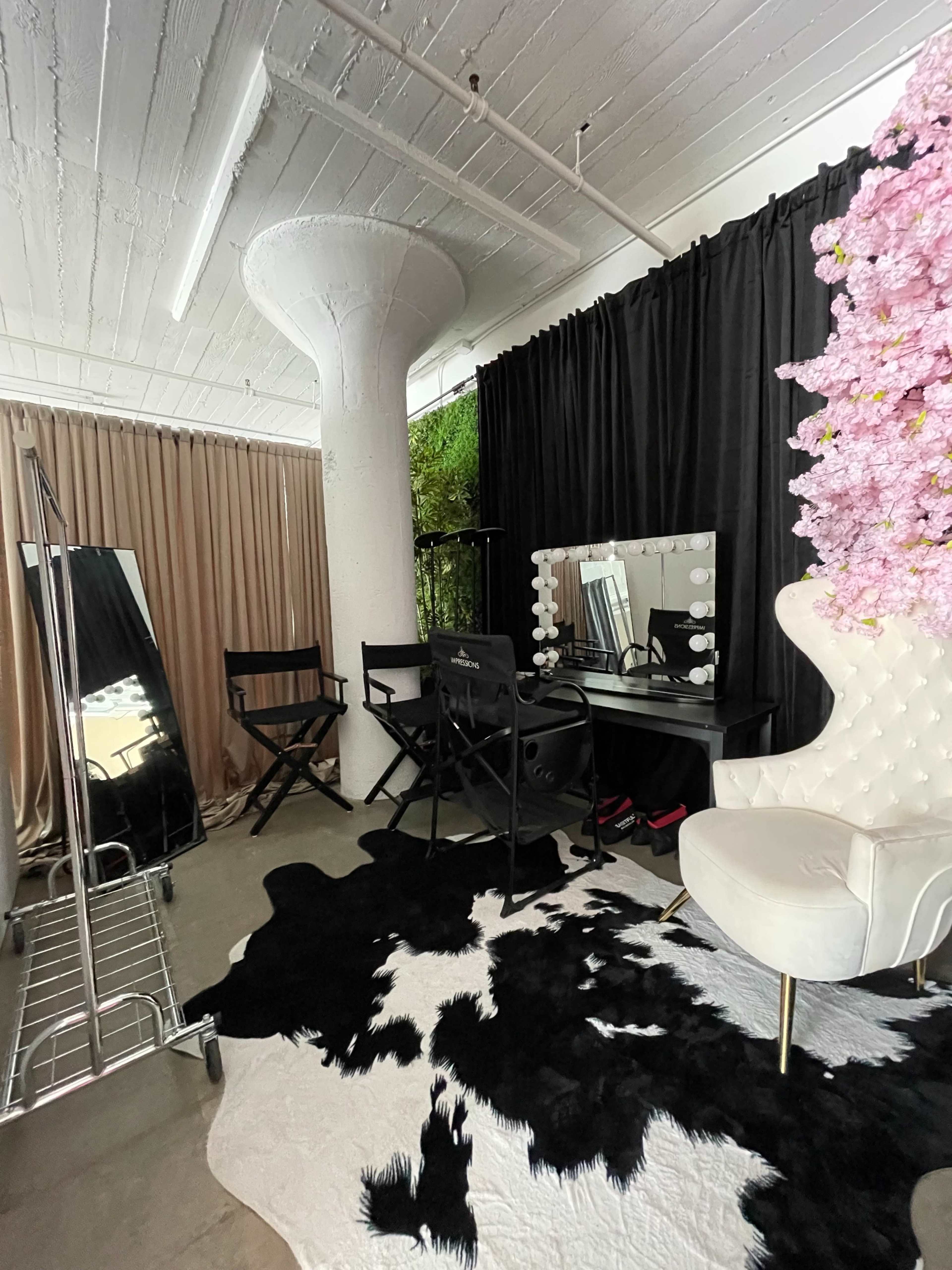 A stylish makeup studio featuring a white chair, a large mirror with lights, a clothing rack, and a cowhide rug underfoot.