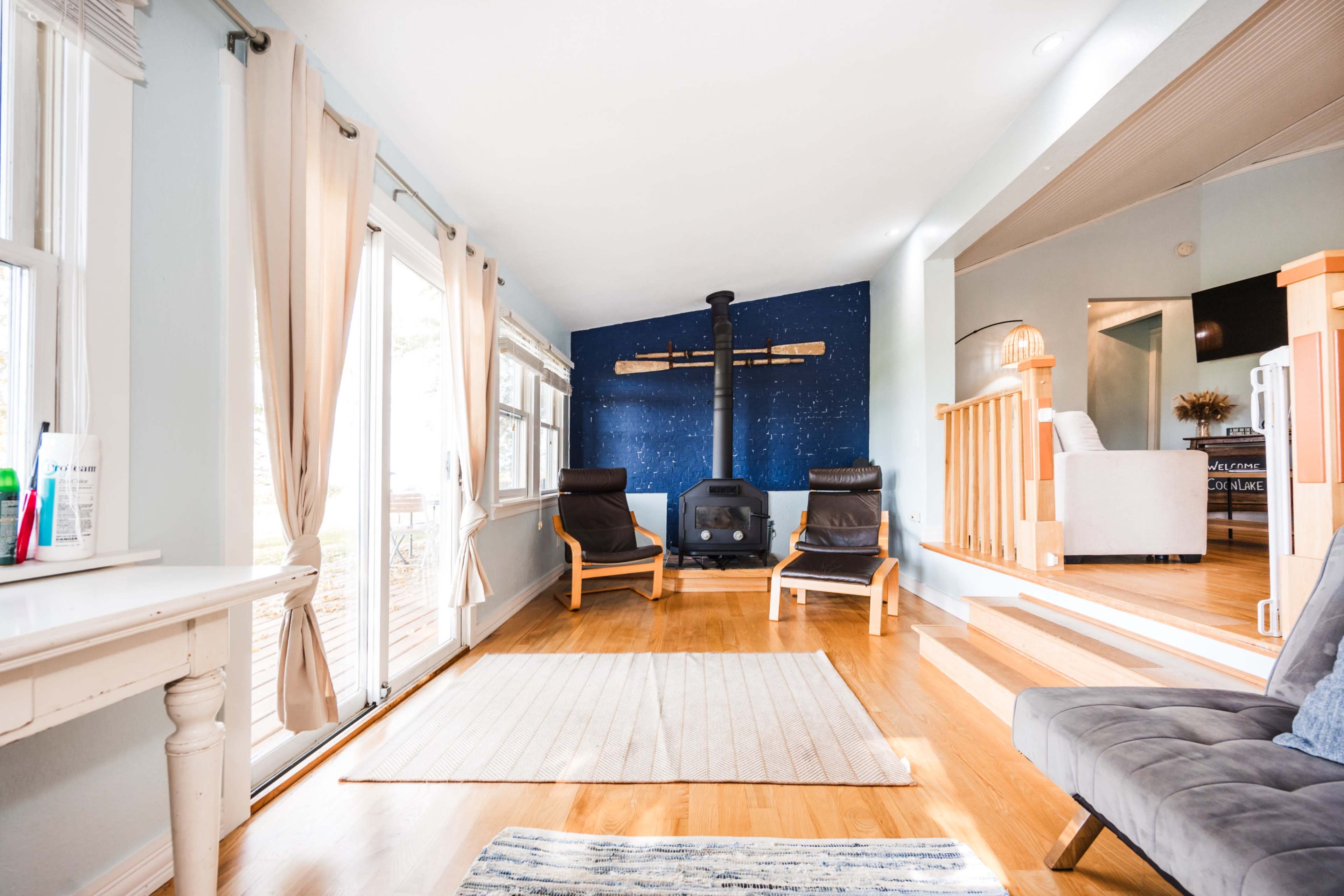 A living room features two black chairs facing a central wood-burning stove against a blue wall, with large windows allowing natural light to enter.
