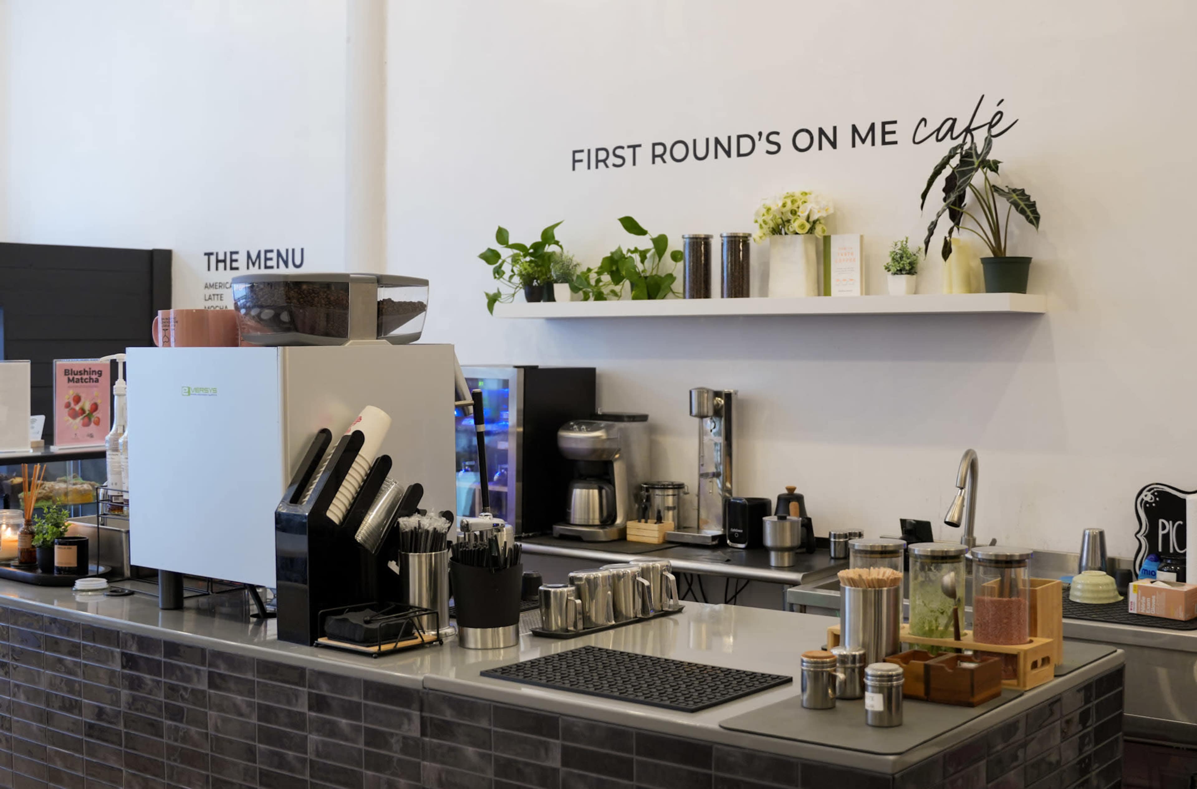 A café counter features various coffee-making equipment, glass jars with ingredients, and a menu, against a backdrop with a decorative phrase about offering a coffee round.