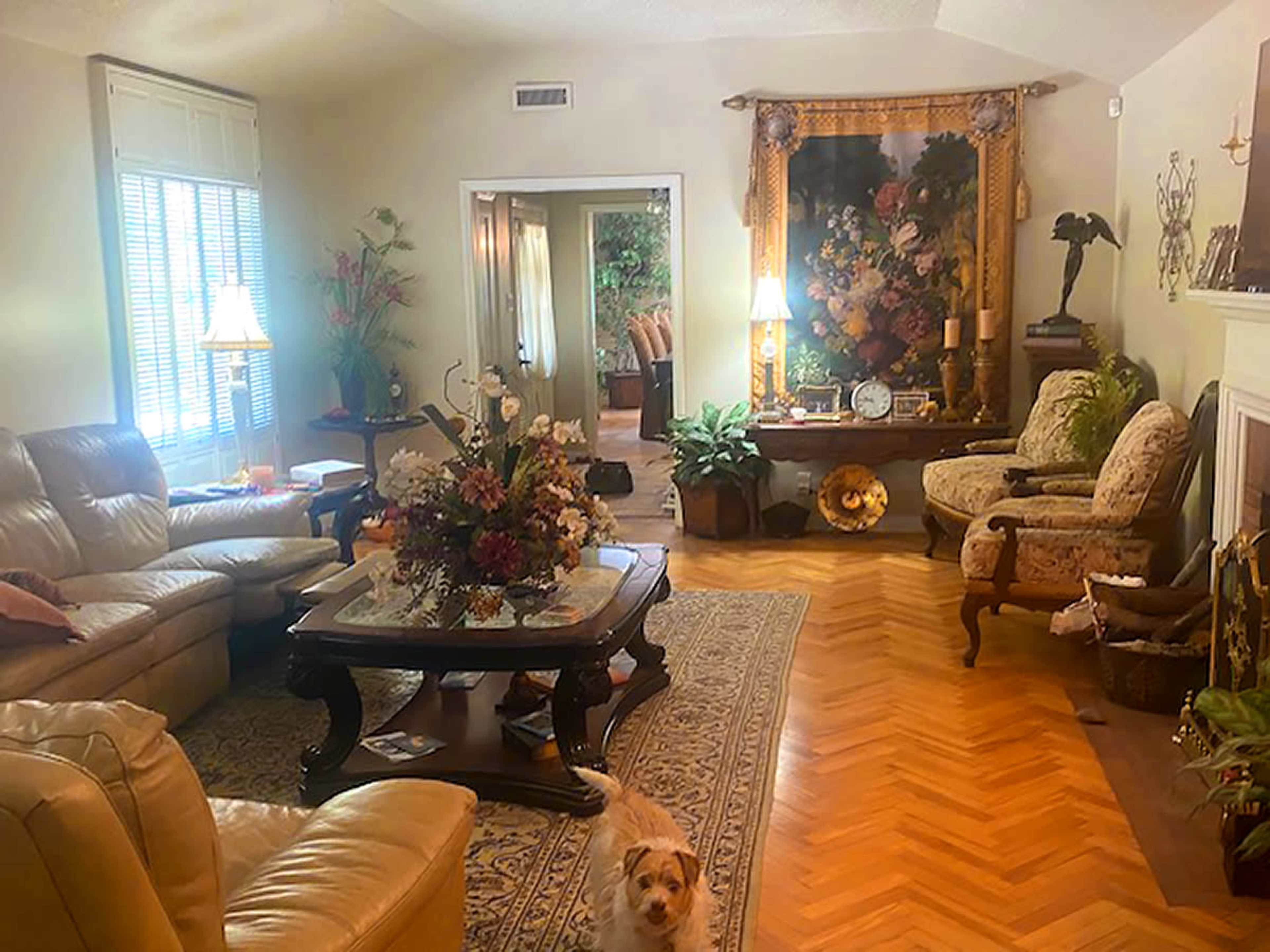 The living room features a mix of leather and upholstered furniture, a floral arrangement on a wooden table, and a patterned rug on a herringbone floor.