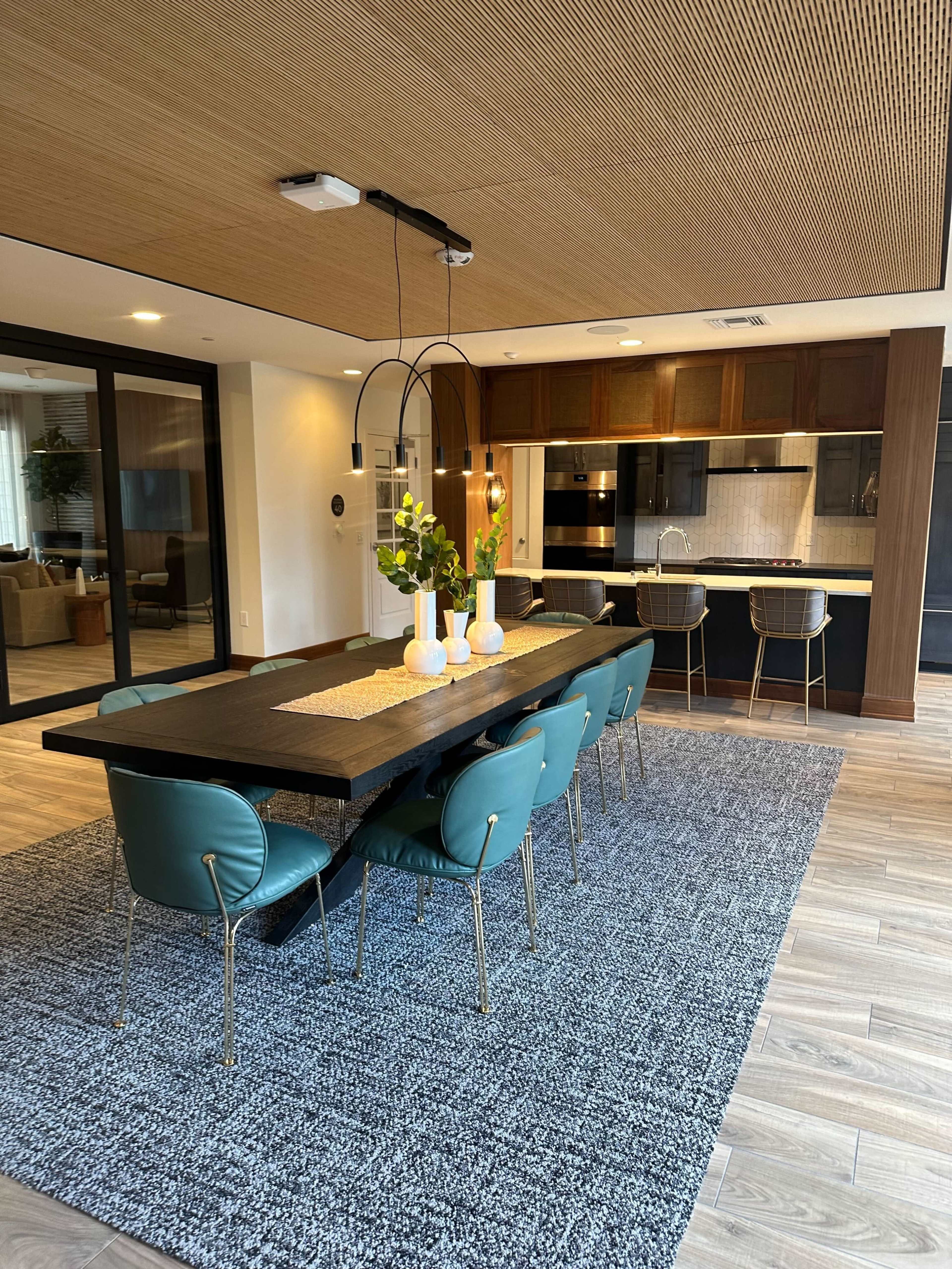 A dining area features a long dark wooden table surrounded by green chairs, with a modern kitchen visible in the background.