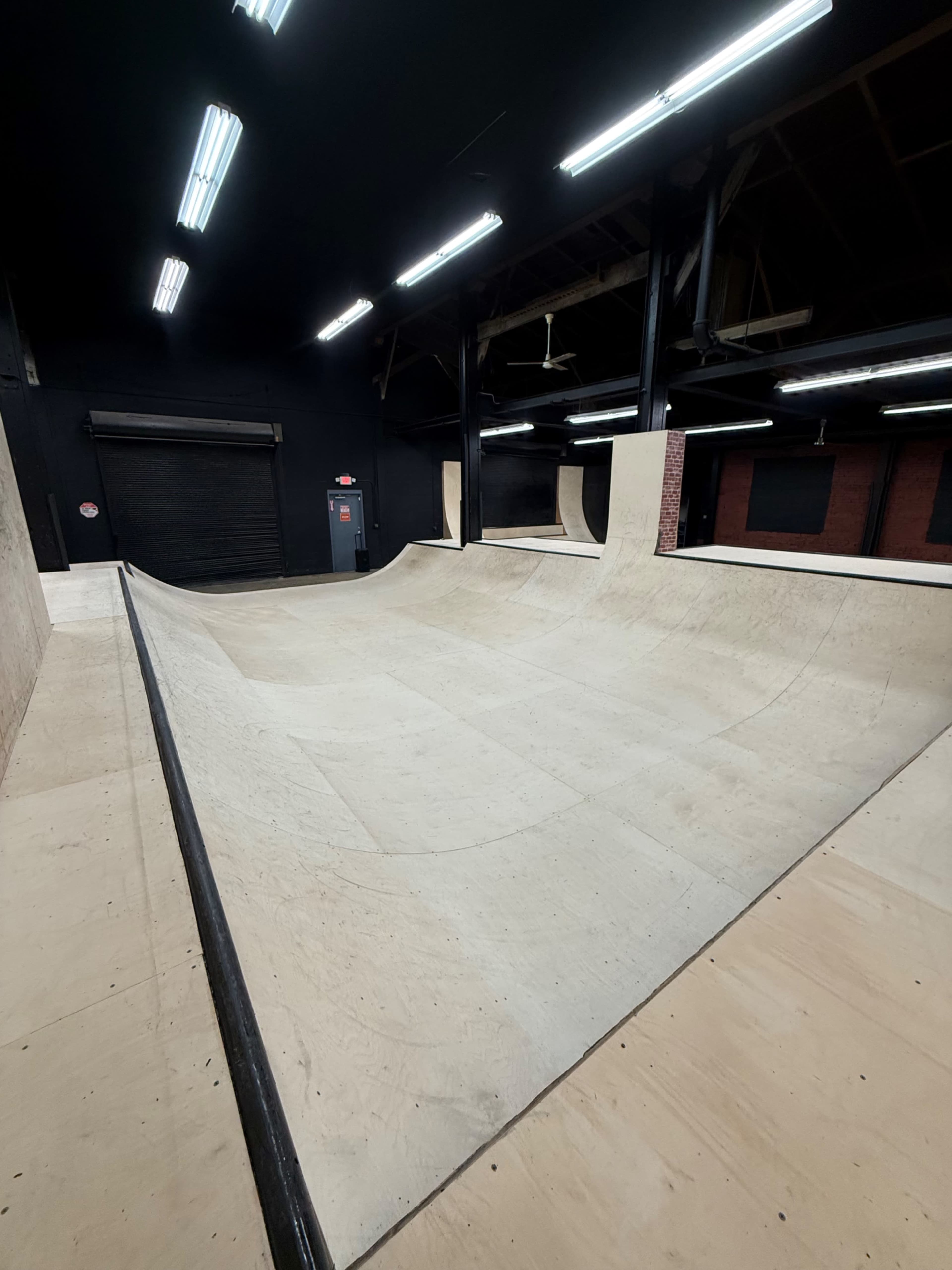 The image shows a spacious indoor skatepark with wooden ramps and a smooth surface, illuminated by overhead fluorescent lights.