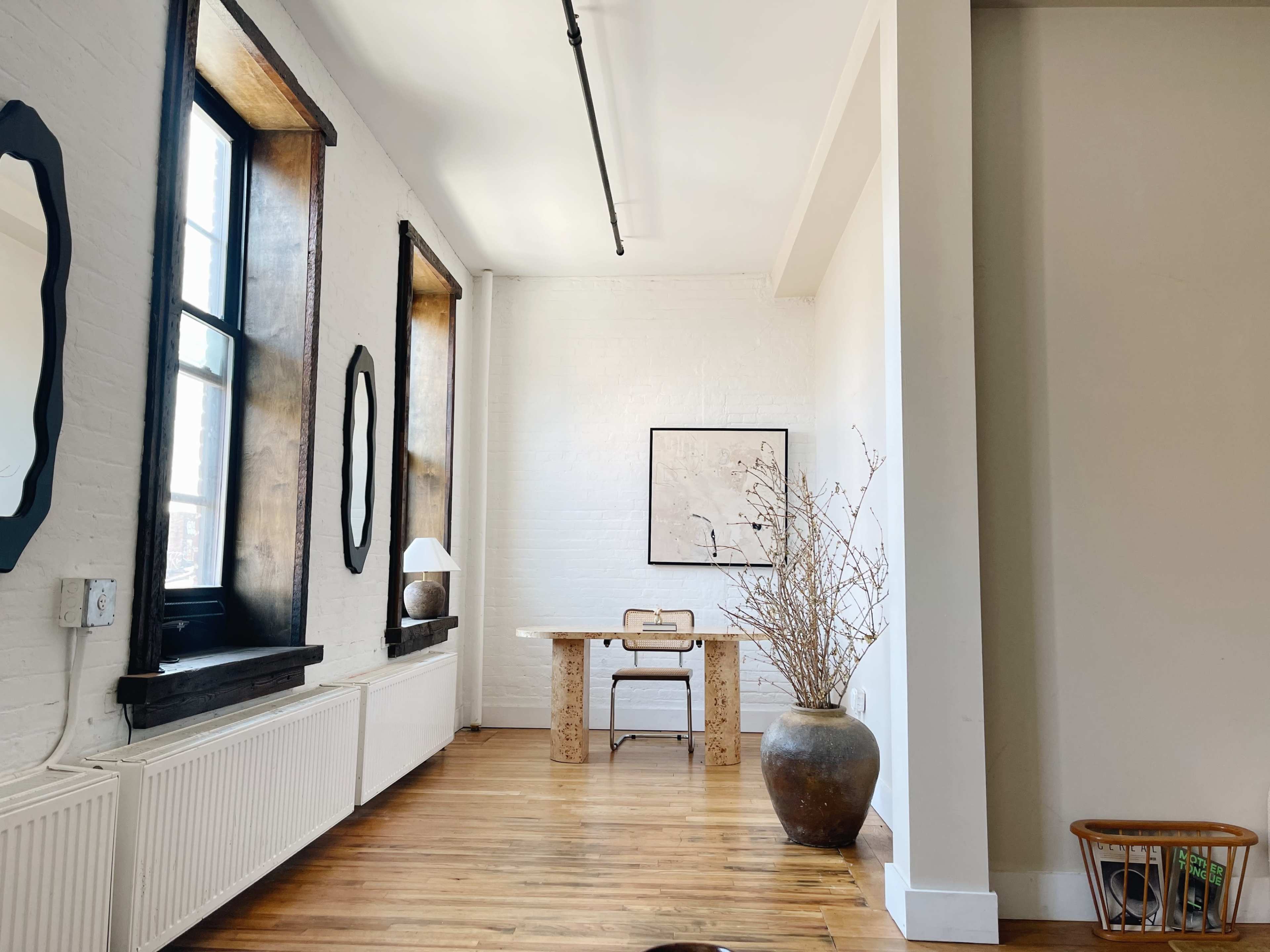 A bright, minimalist room with large windows, a wooden table, a chair, and a decorative vase containing branches.