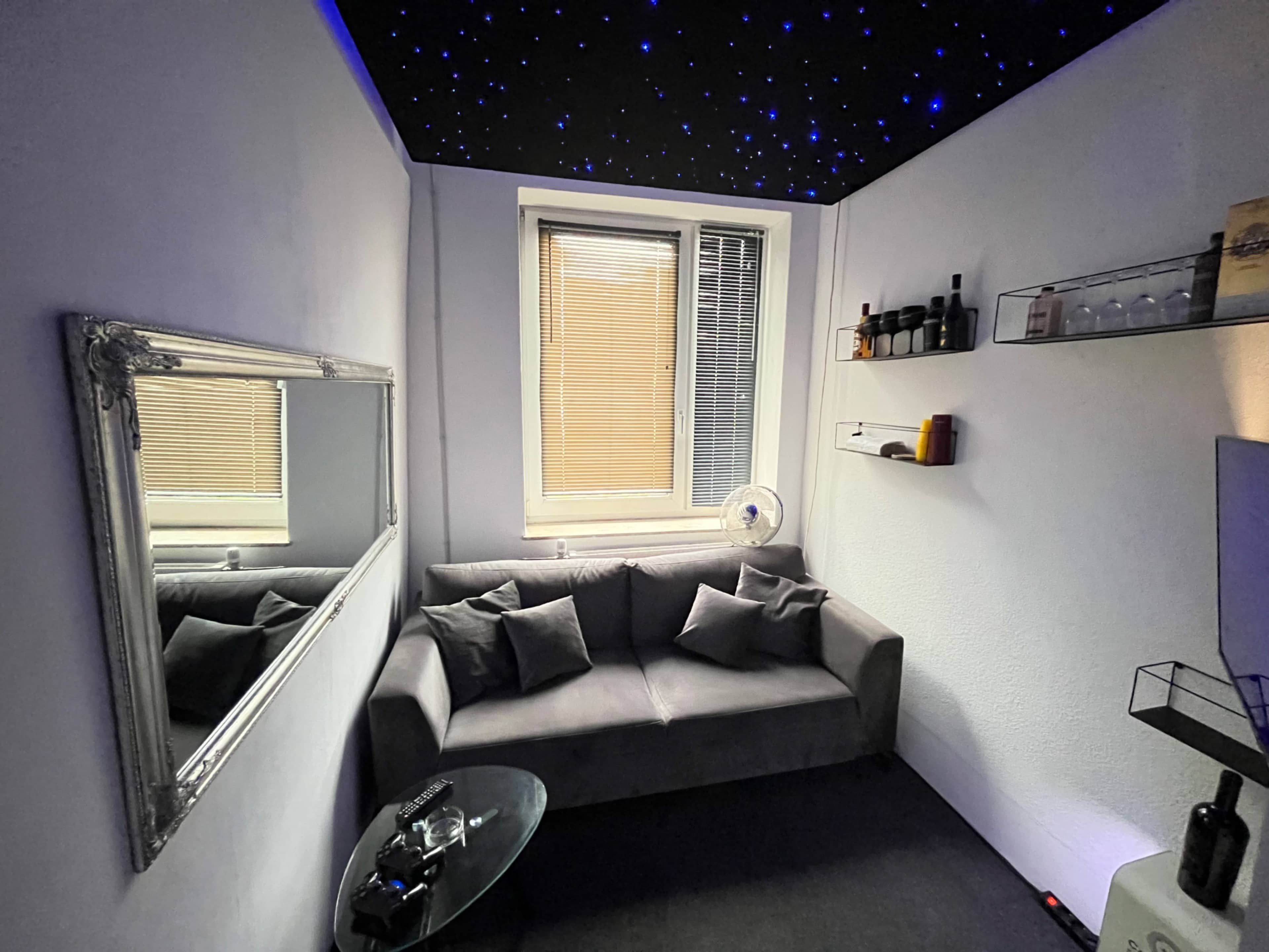 A small room with a gray sofa, a glass coffee table, a mirror on the wall, and shelves displaying several bottles.