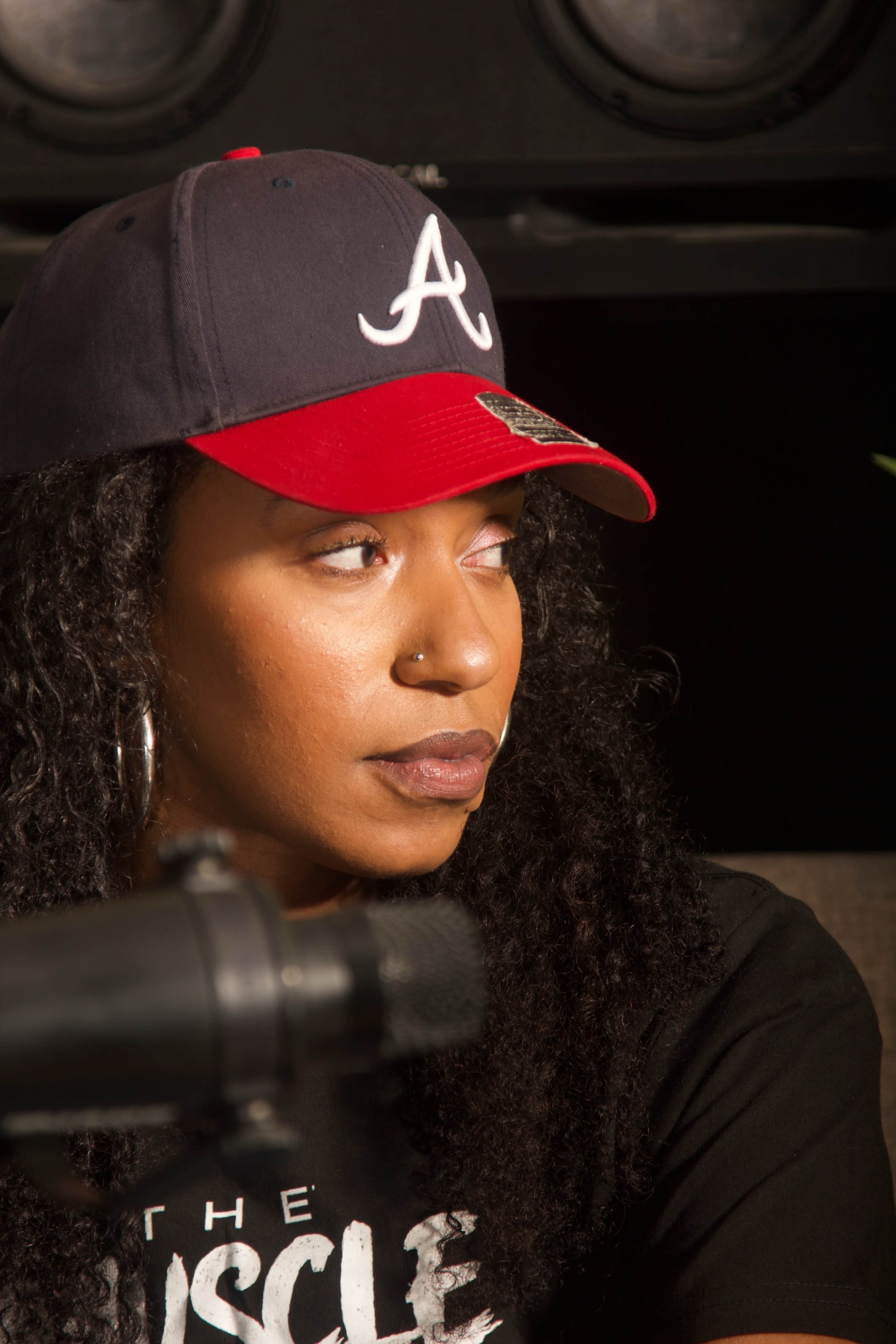 A woman with curly hair wearing a red and navy baseball cap and a black t-shirt is seated in front of a microphone, looking thoughtfully to the side.