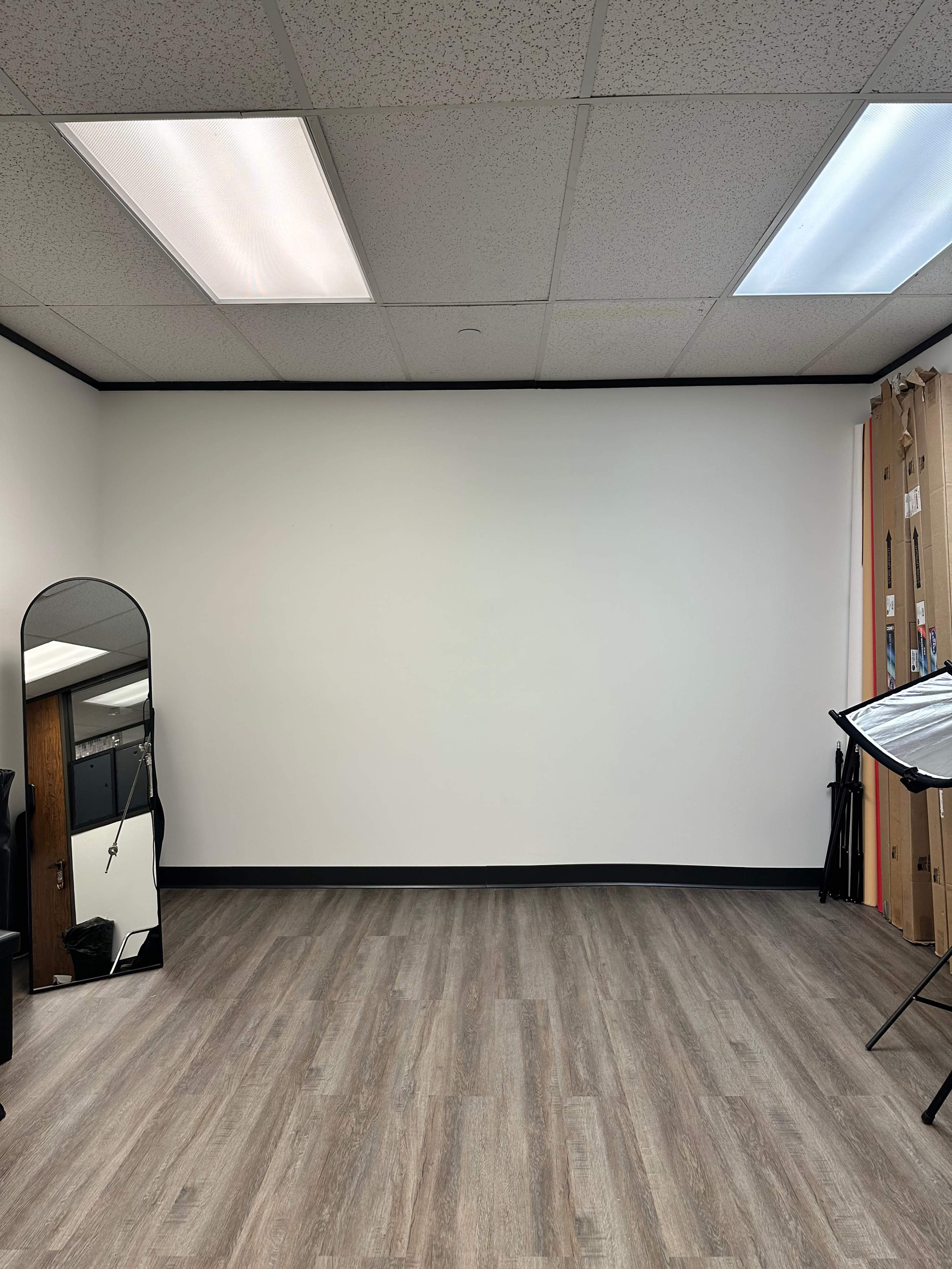 A blank, well-lit room with a large mirror on one side and studio equipment arranged along the walls.