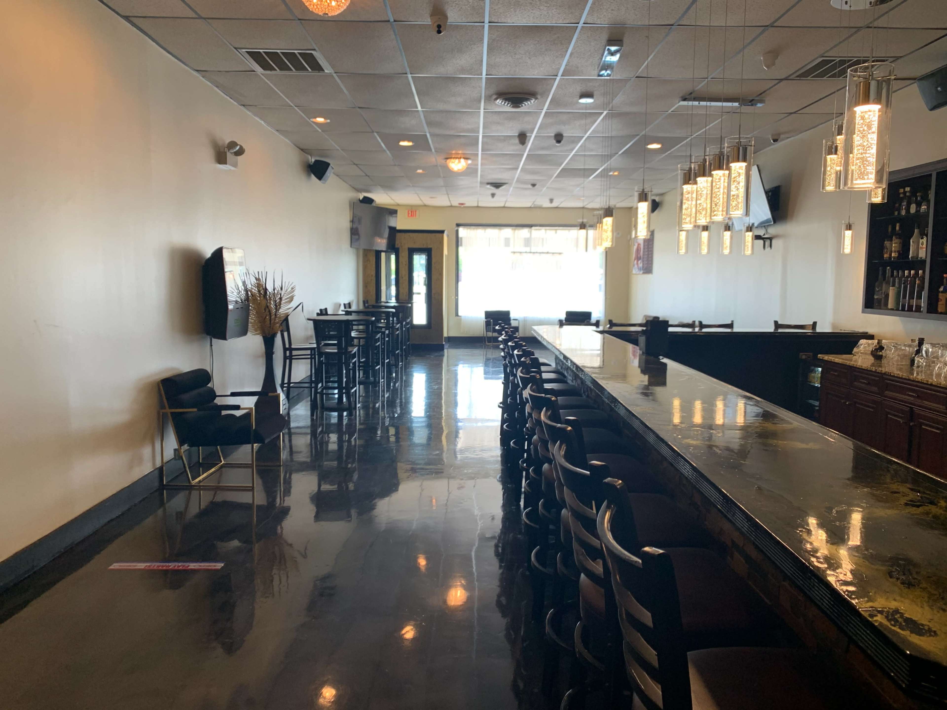 A well-lit bar area with a long counter, high-backed stools, and sparse seating along the walls.