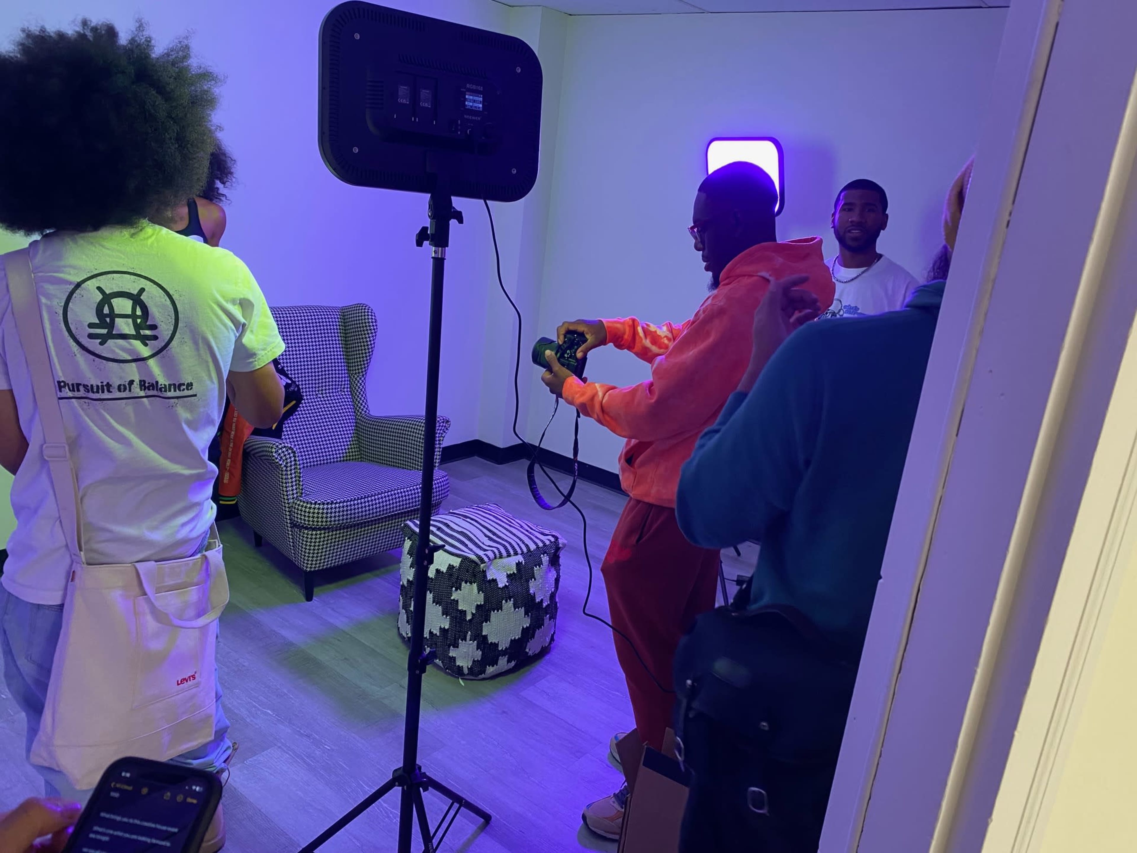 A group of people is gathered in a room with purple lighting, where one individual is holding a gaming controller while others appear to be engaged in conversation.