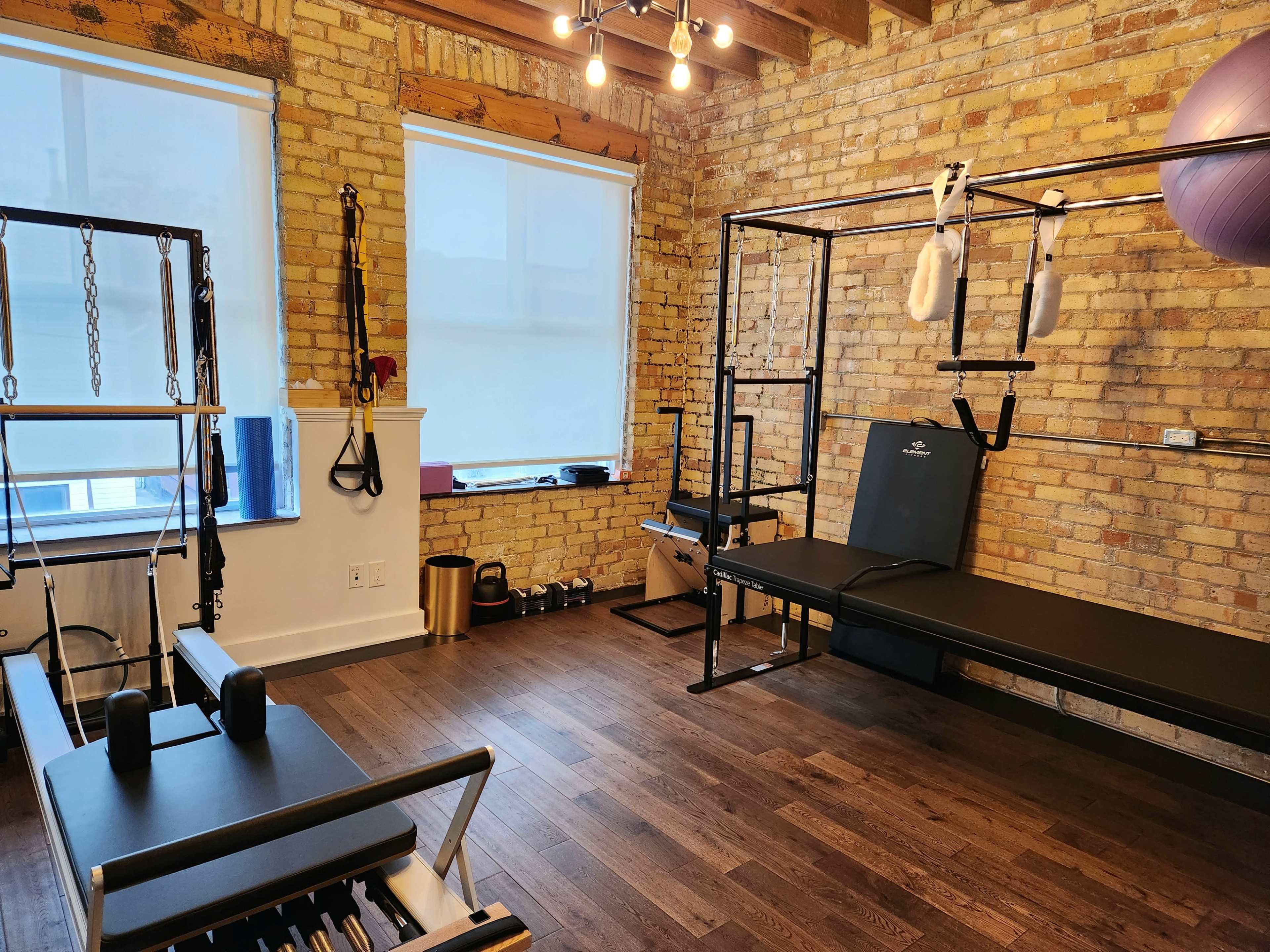 A fitness studio with wooden flooring, exposed brick walls, and various workout equipment, including a reformer, a bench, and fitness accessories.