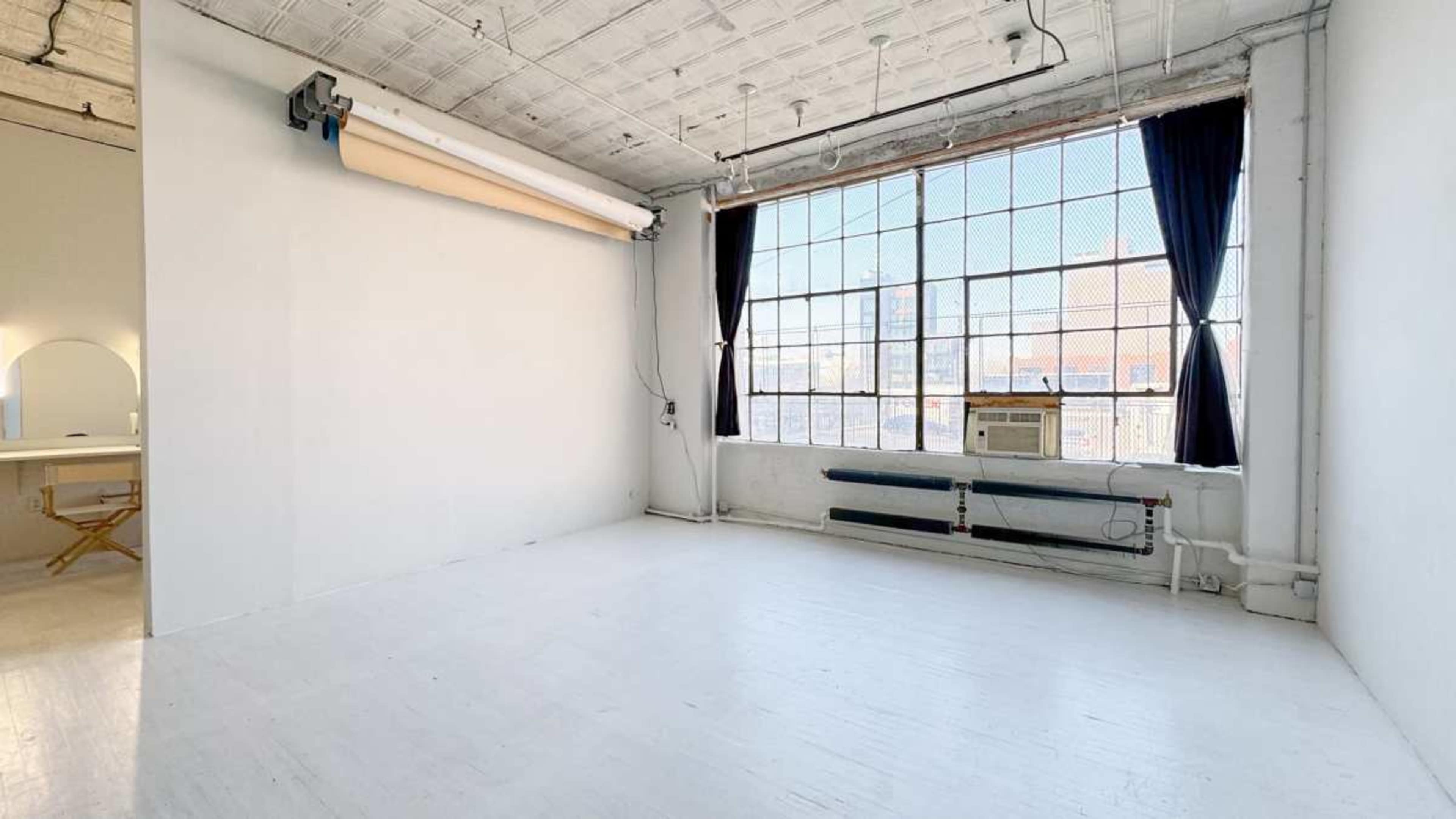 The image shows a bright, empty studio space with large windows and white walls.