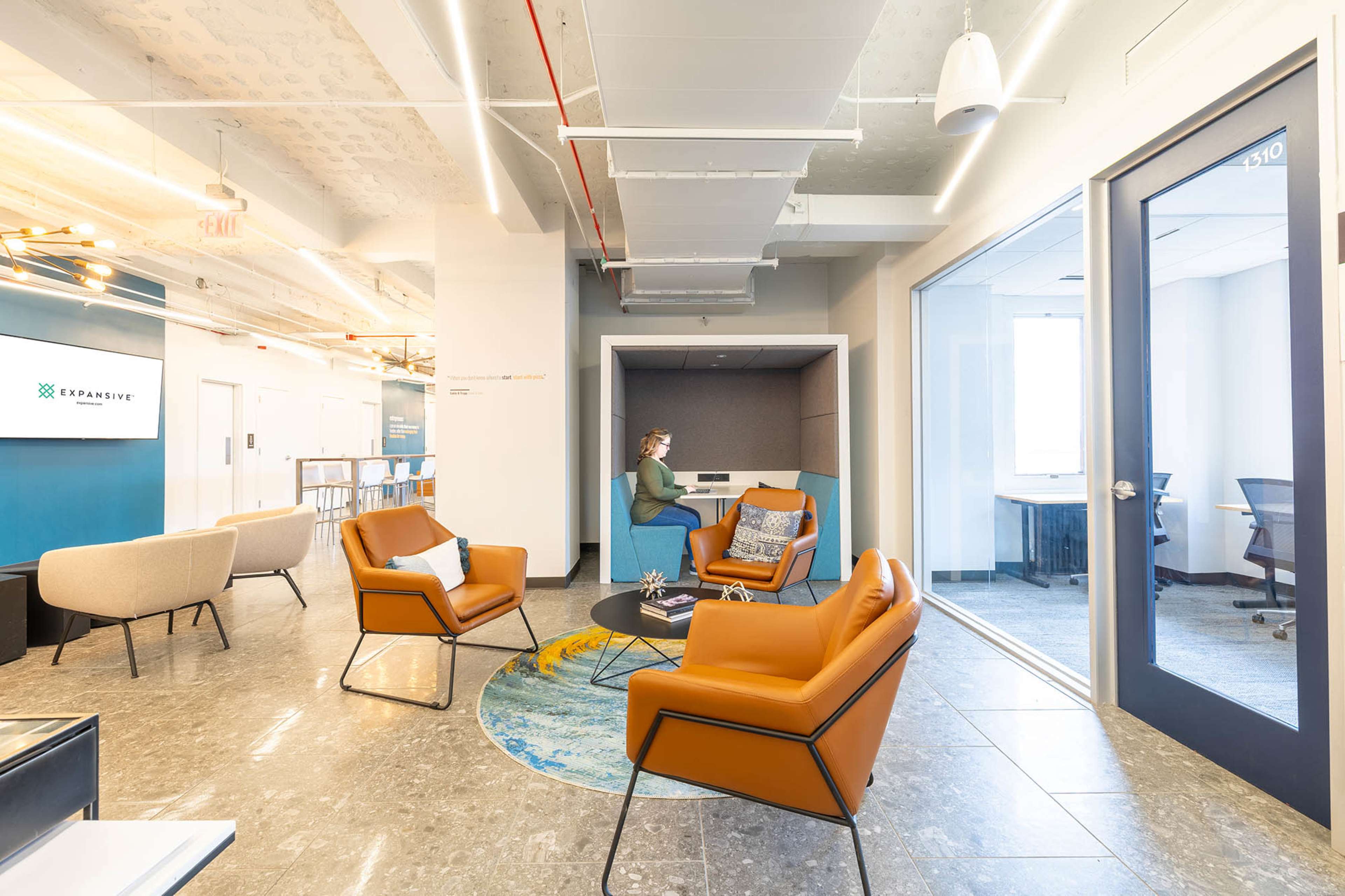 The image shows a modern office lounge area featuring orange chairs, a round rug, and a glass-walled meeting room.