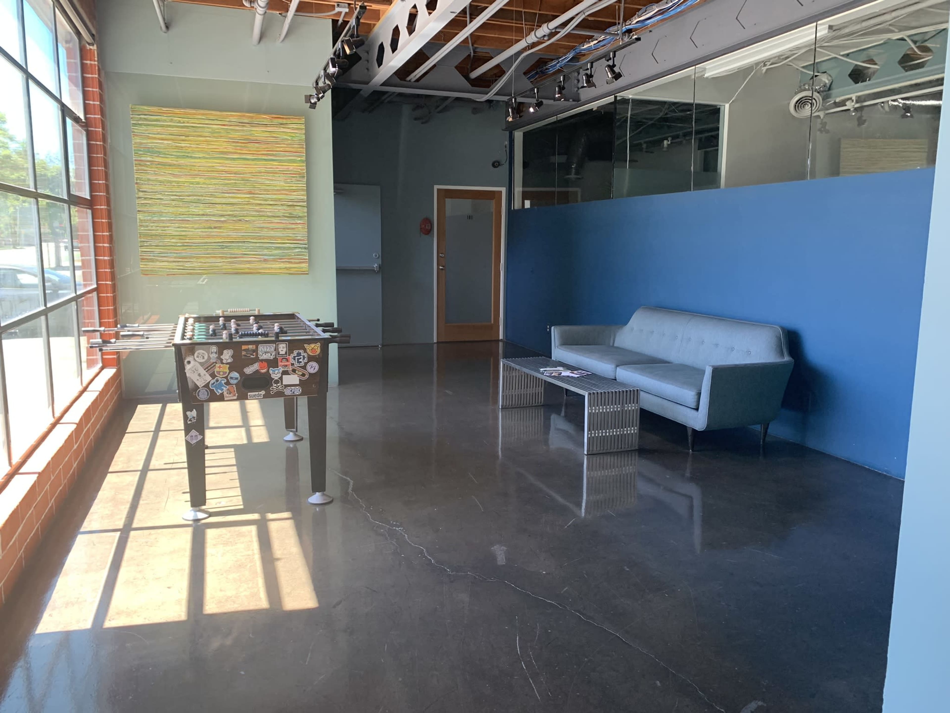 An empty room with a modern gray couch, a foosball table decorated with stickers, and large windows allowing natural light to enter.