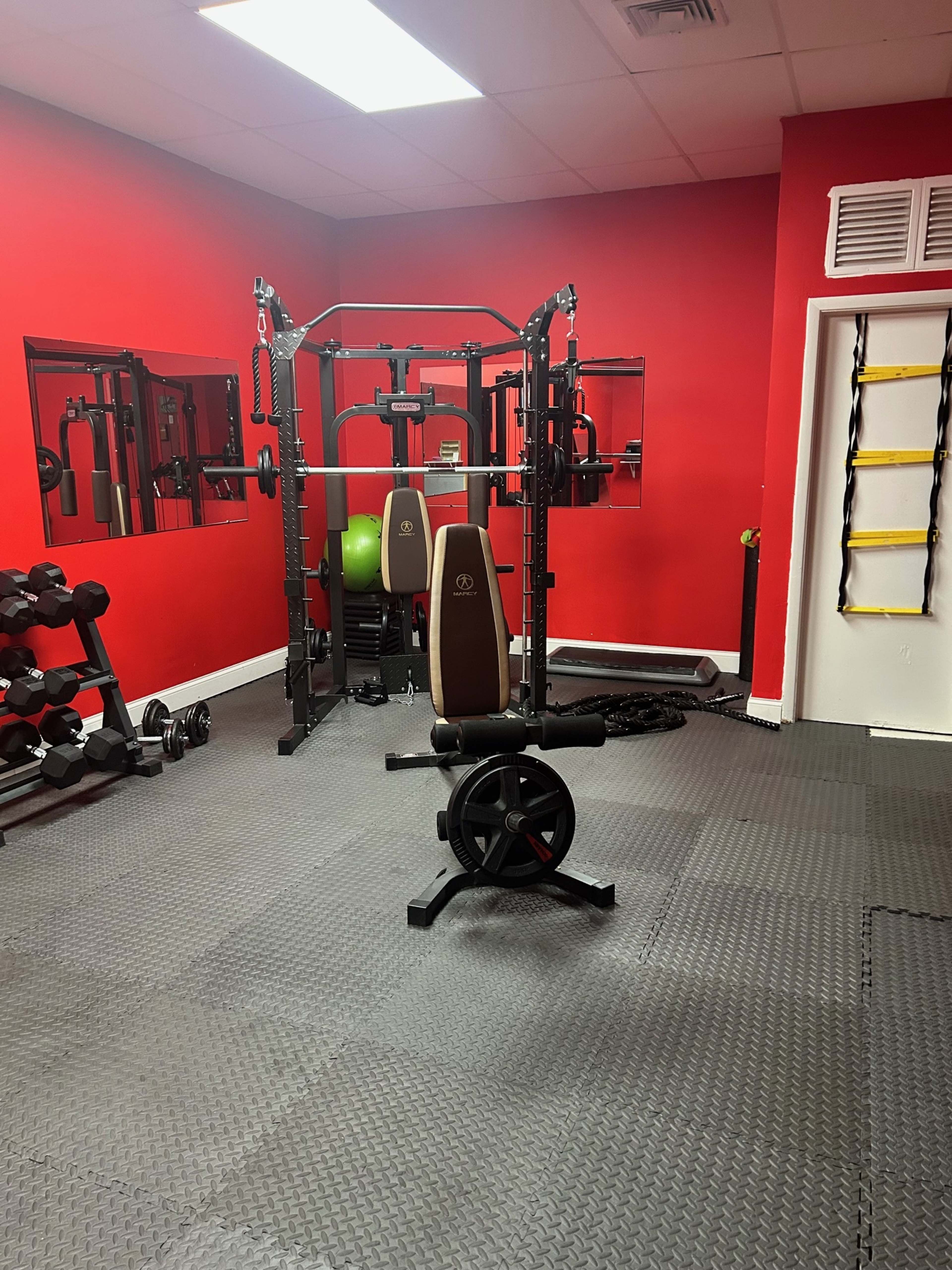 The image shows a gym interior with red walls, various exercise equipment including weights, a multi-station machine, and a mat flooring.