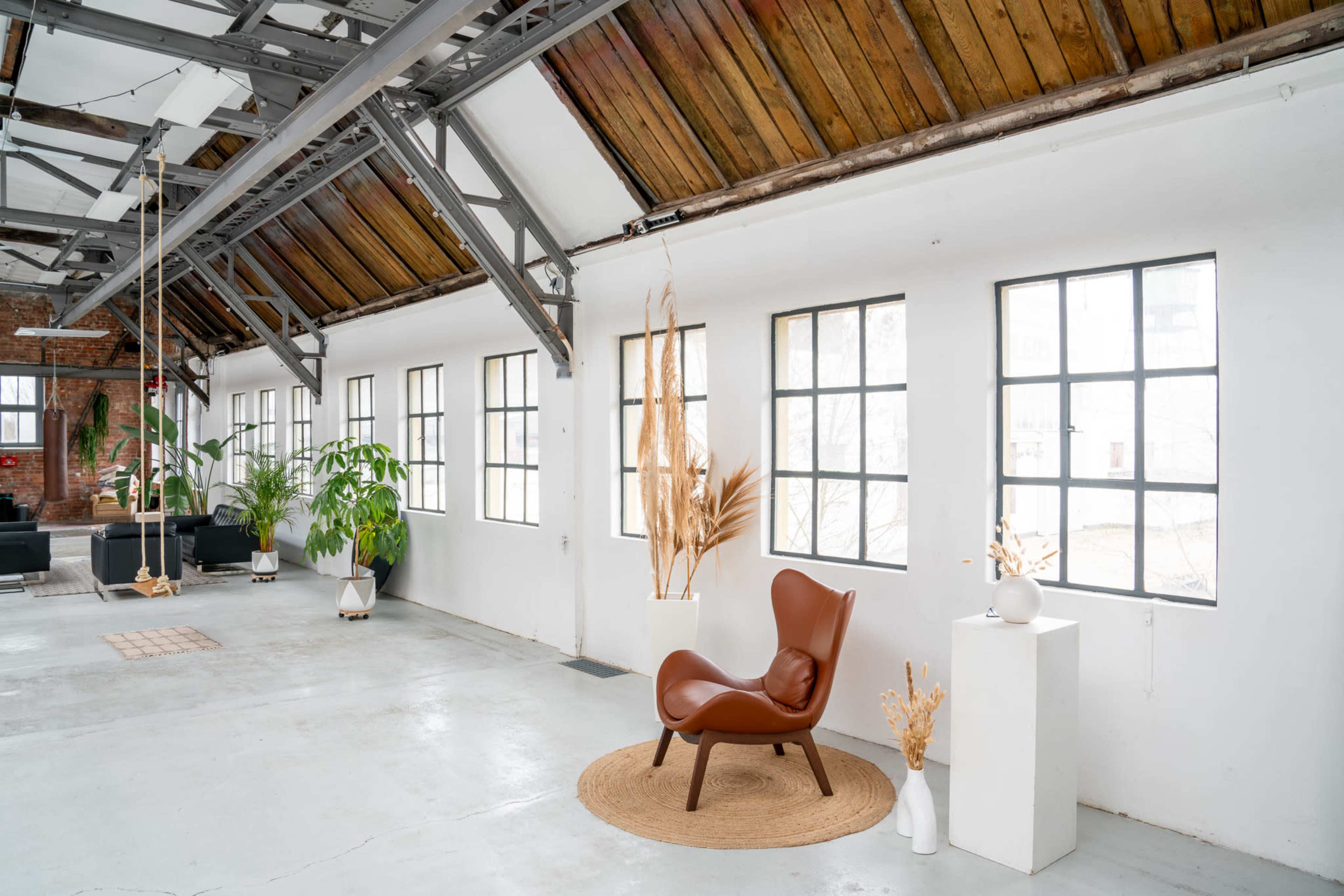 A spacious, industrial-style room with large windows, featuring a brown chair on a round rug and decorative plants.