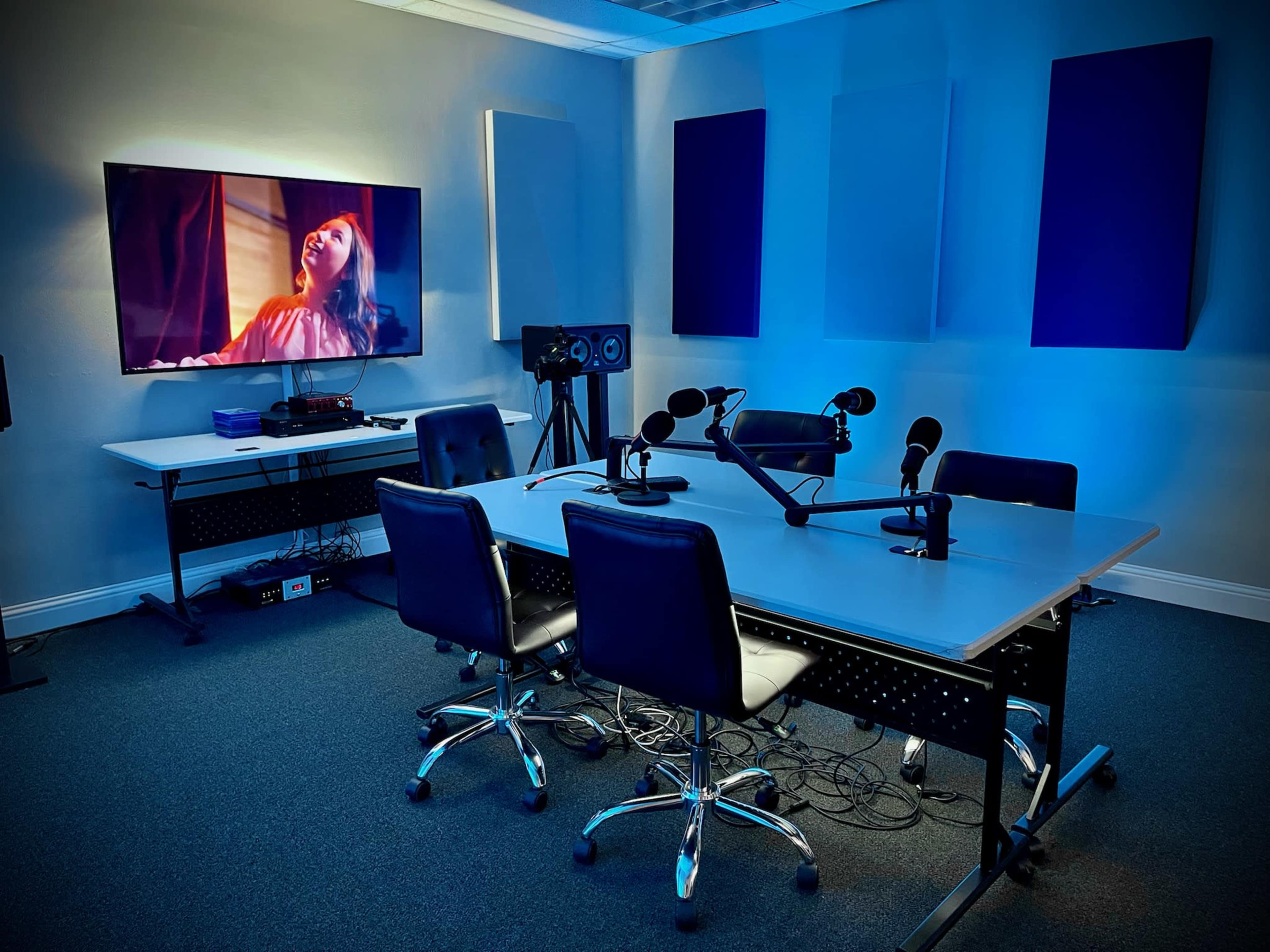 A modern recording studio featuring a large screen, several microphones, and a table with chairs arranged for a podcast or recording session.