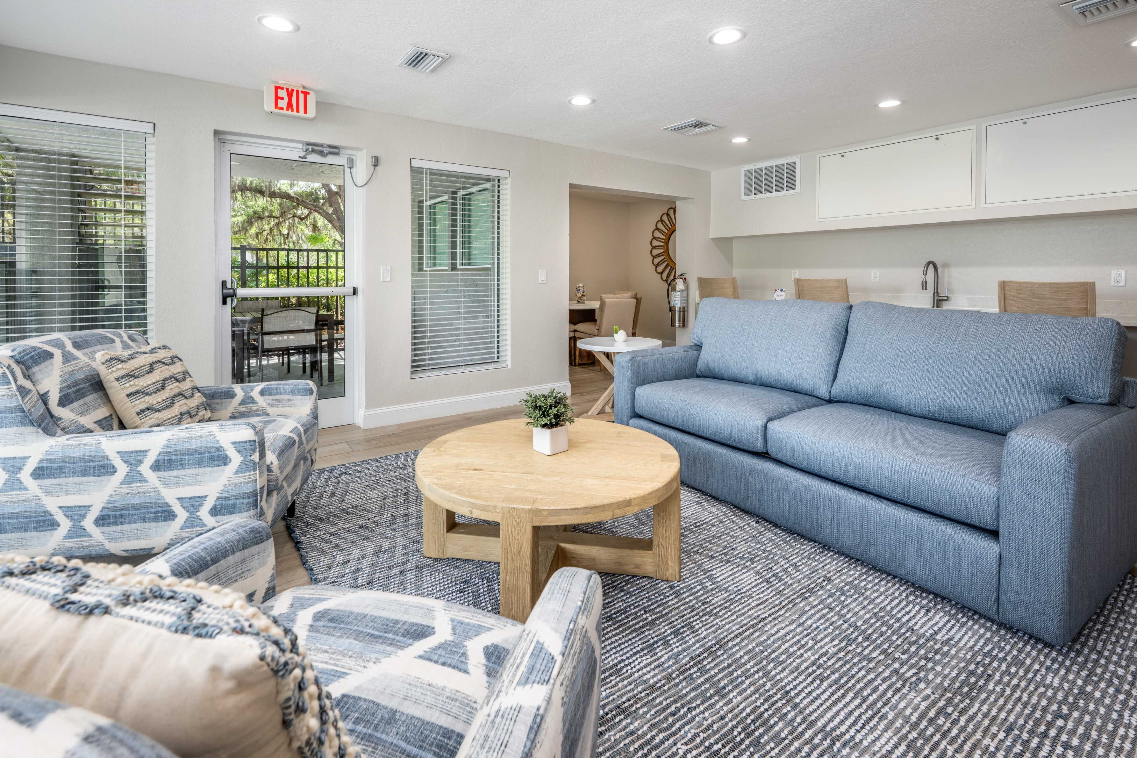The image shows a modern living area furnished with a blue sofa, two patterned armchairs, and a round wooden coffee table, all set against a backdrop of large windows and a light-colored interior.