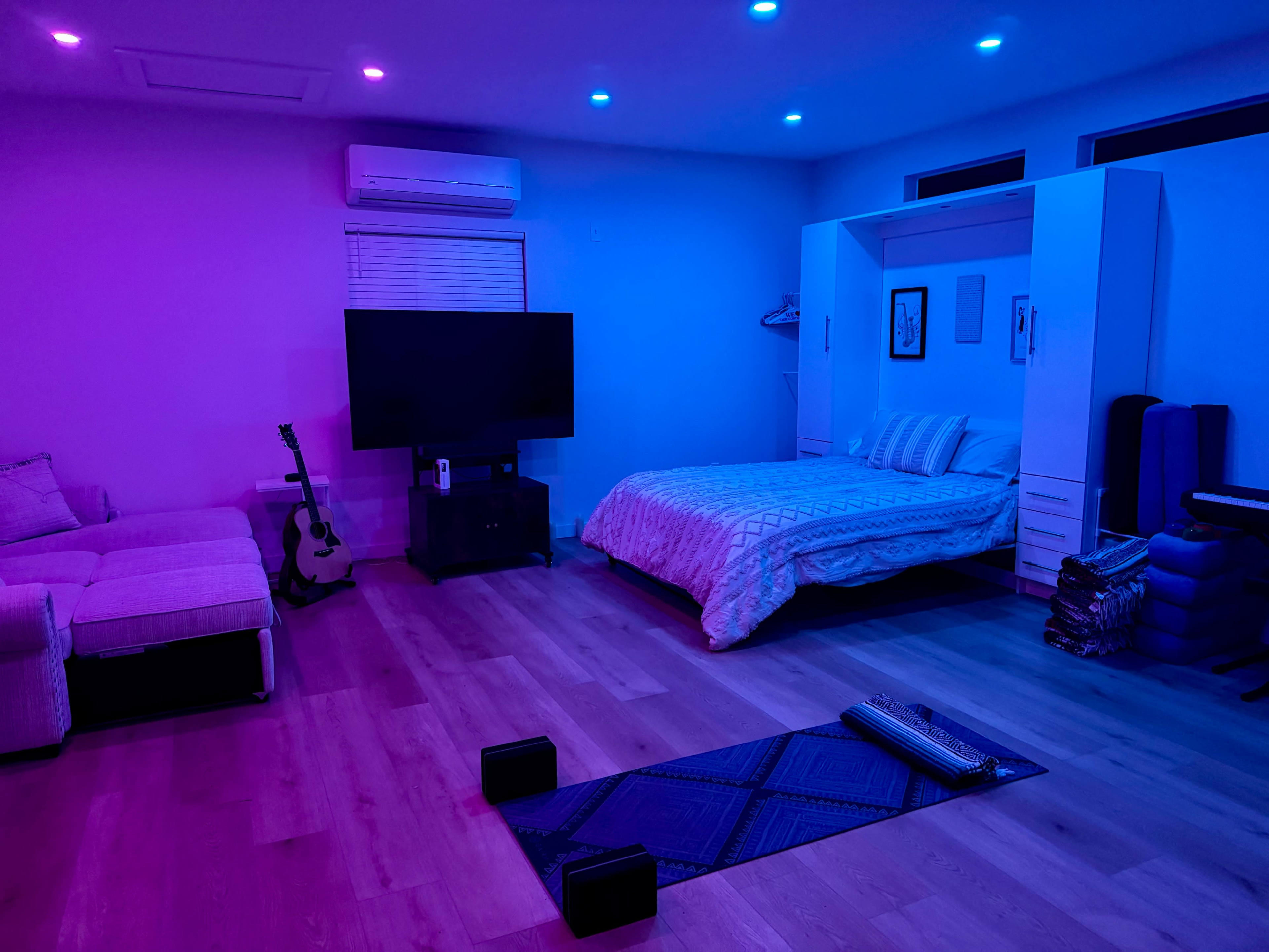 The room features a bed with light-colored bedding, a large television, a couch, and exercise equipment, all illuminated by purple and blue lights.