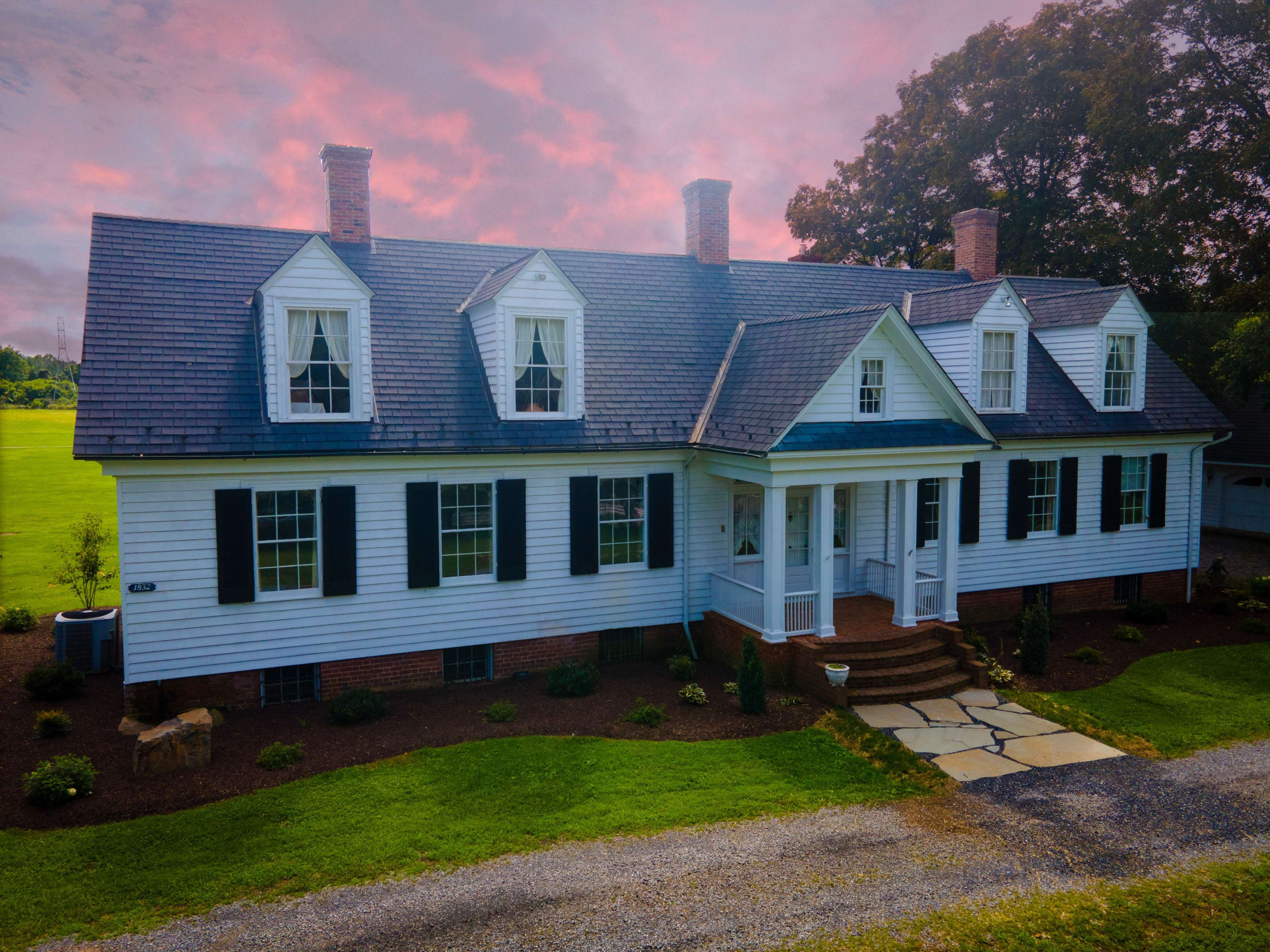 A two-story white colonial-style house features a symmetrical facade, with dormer windows and a front porch, set against a backdrop of a colorful sky.