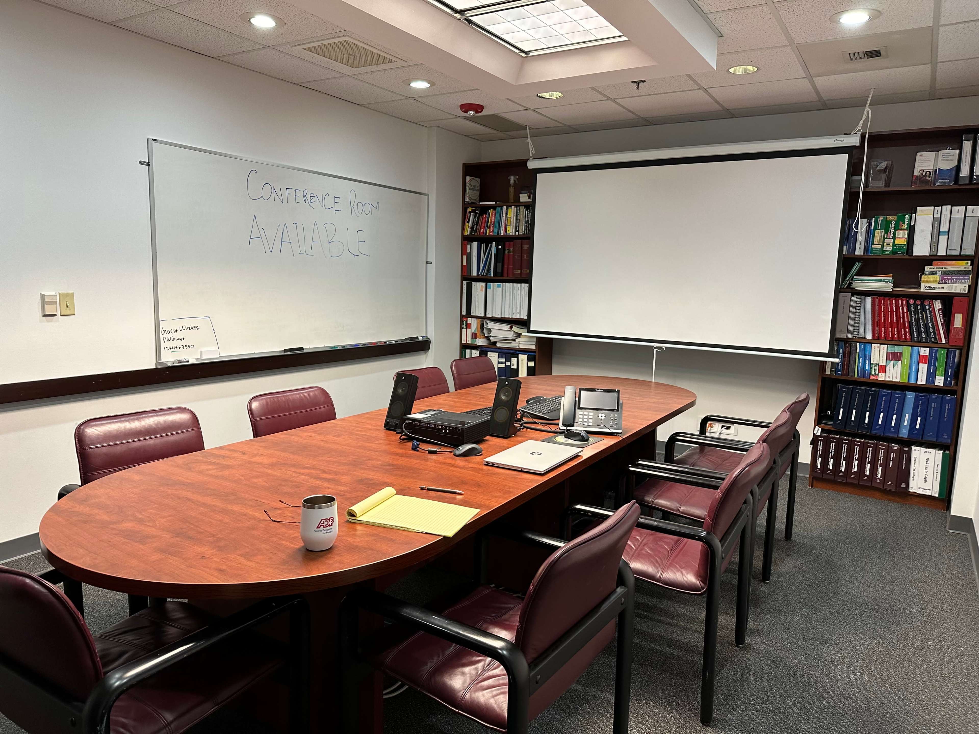 A conference room is set up with a large wooden table, chairs, a projector screen, a whiteboard, and office equipment.