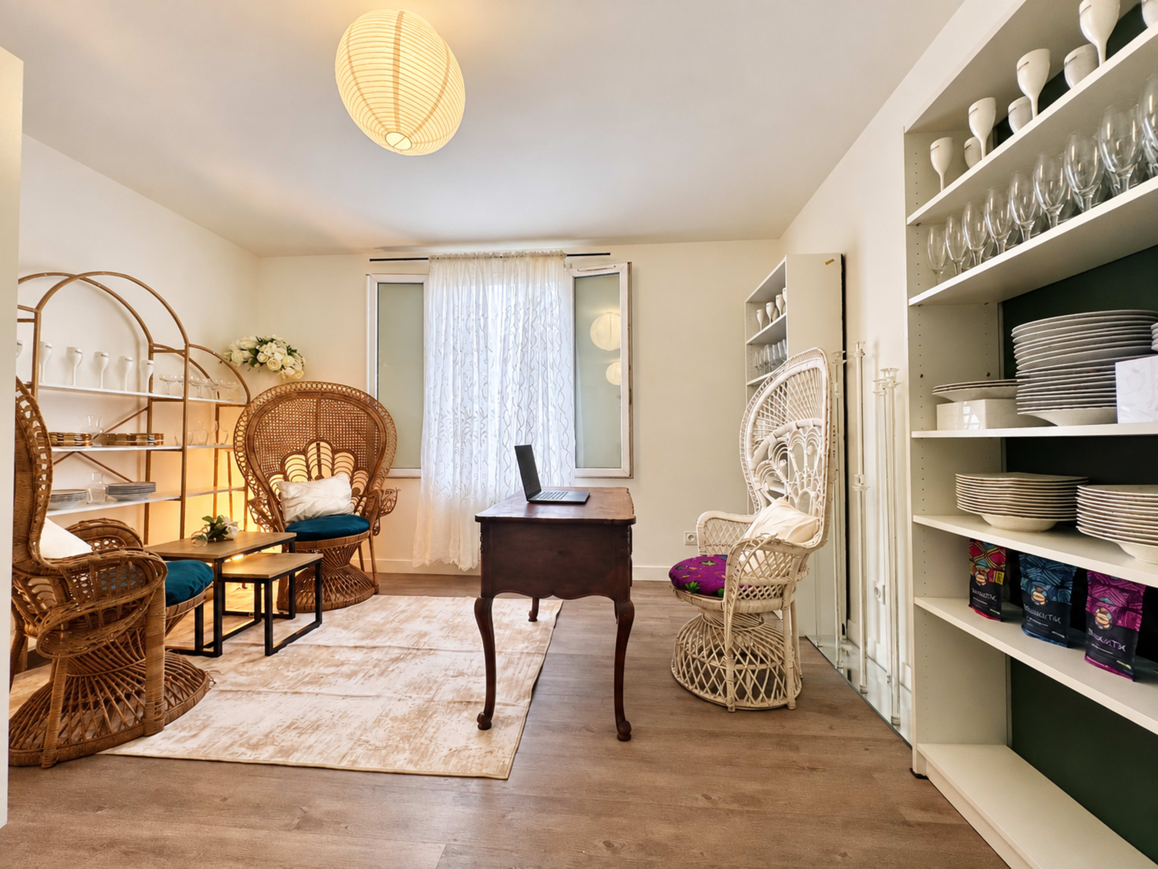 The image features a cozy interior space with a wooden desk, two wicker chairs, a shelf of dishes, and large windows letting in natural light.