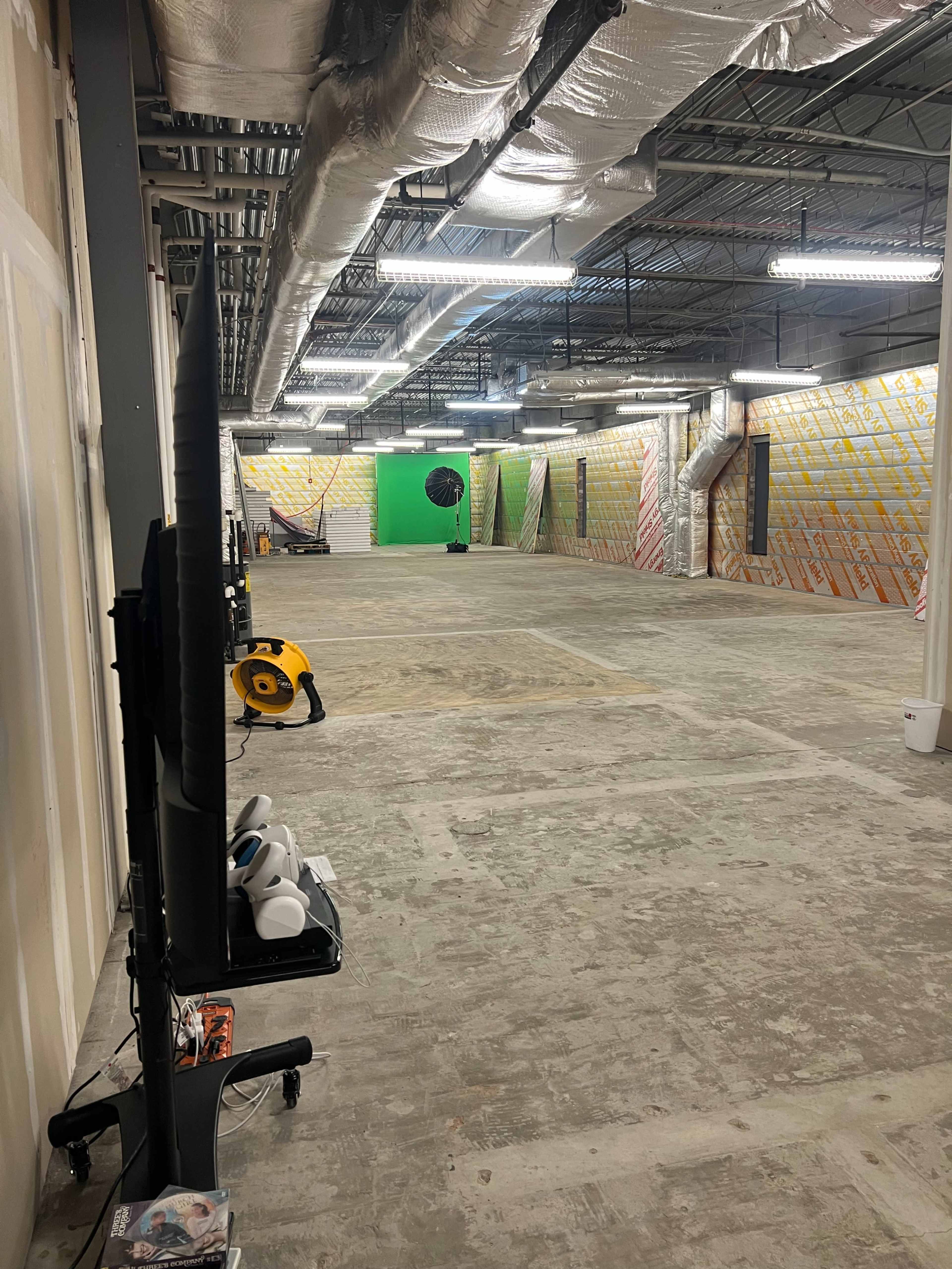 The image shows an empty warehouse-like space with exposed ventilation, a green screen at one end, and various equipment scattered around.