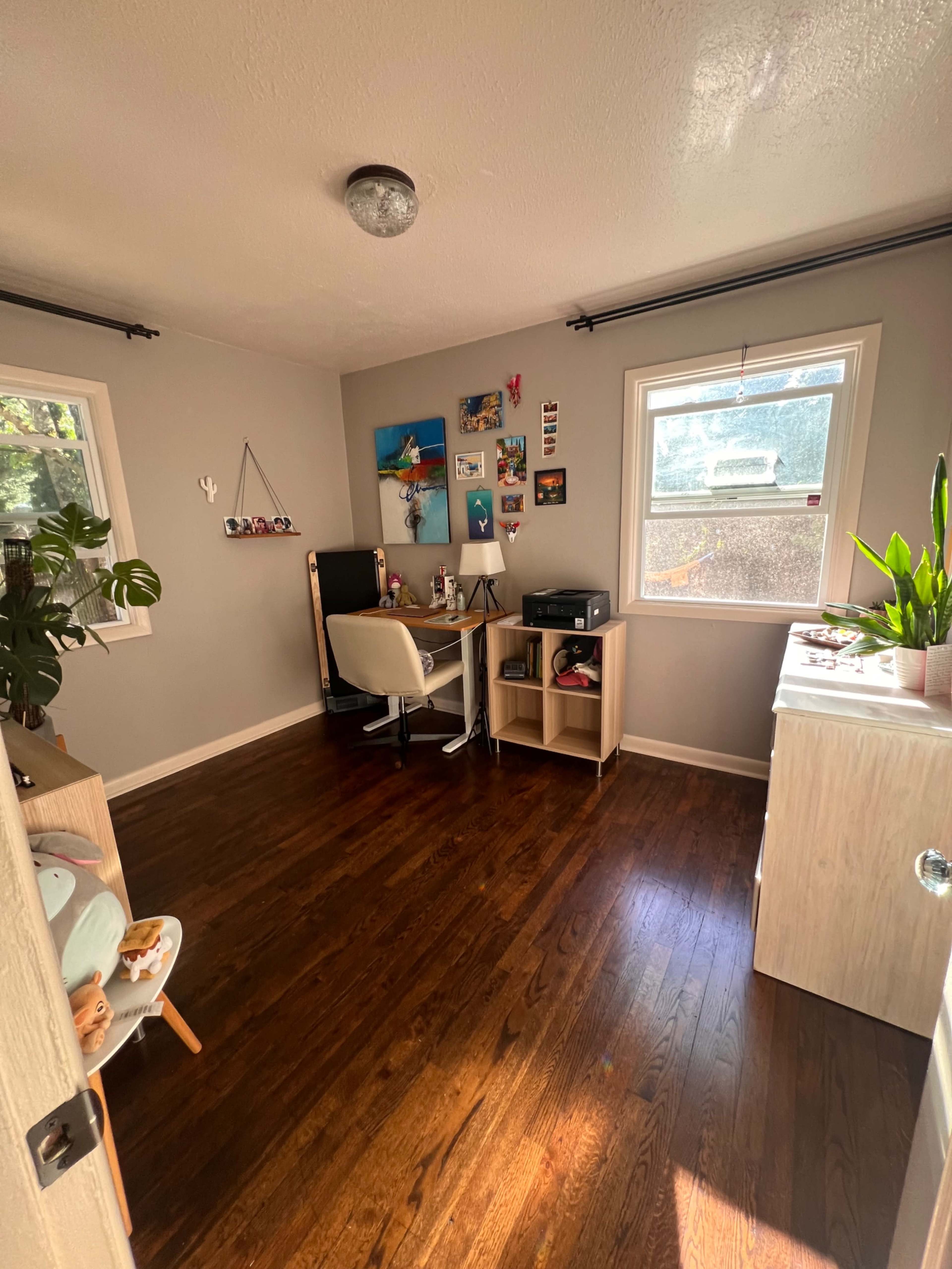 The image shows a small room featuring a desk with a chair, a bookshelf, and a window with plants on the sill.