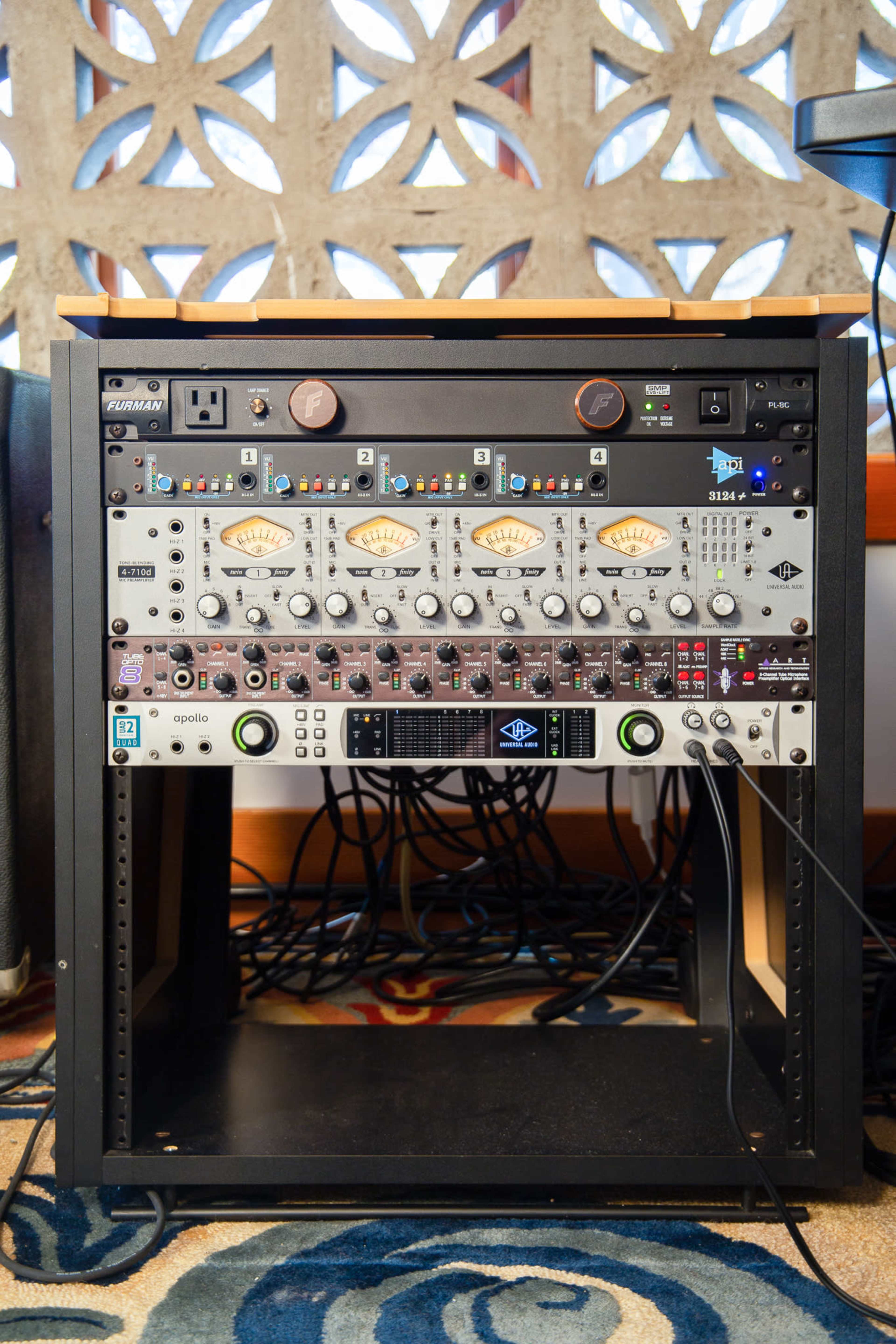 A professional audio rack with several pieces of equipment, including processors and audio interfaces, positioned beneath a tabletop and surrounded by cables.