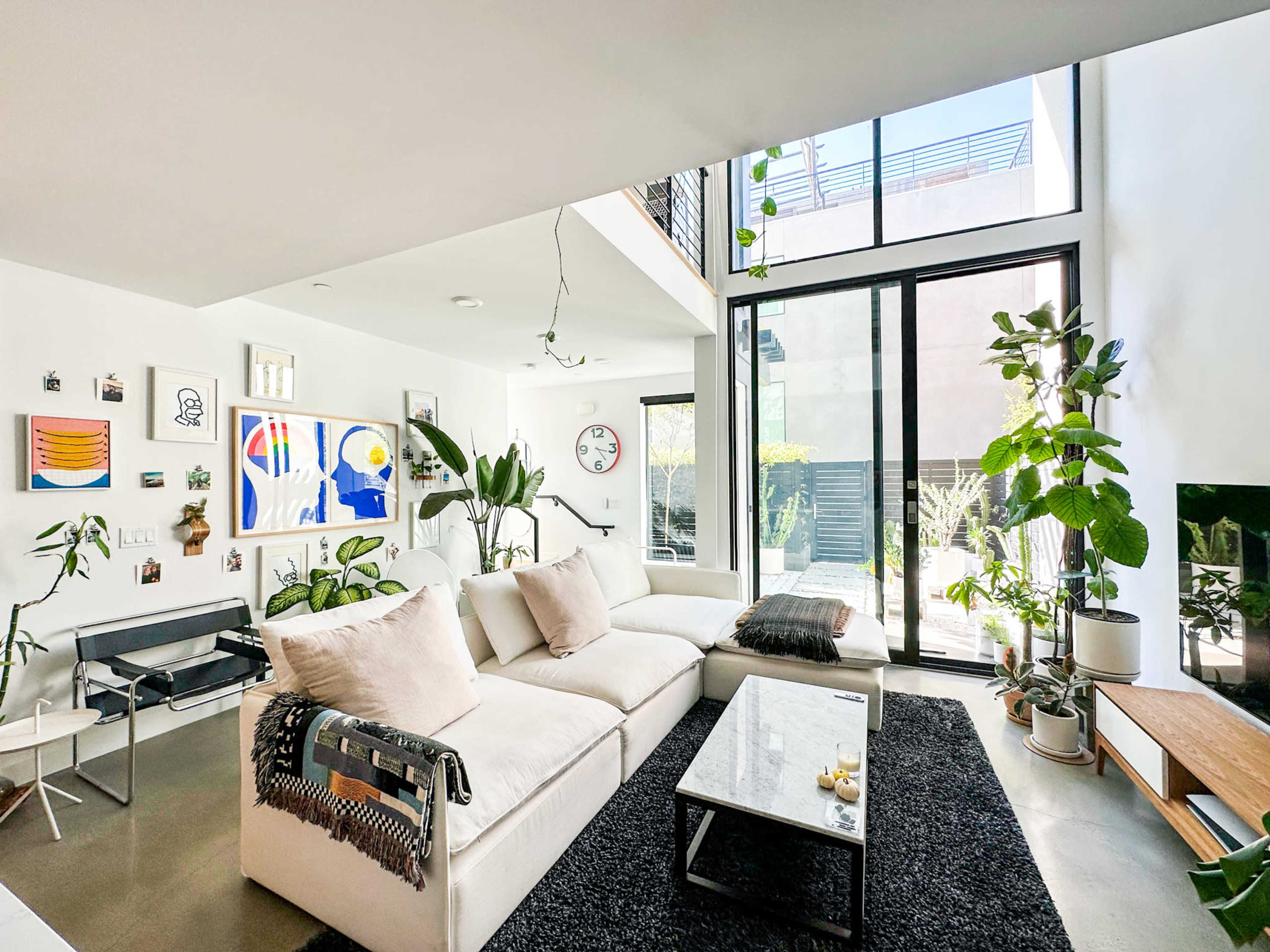 The image shows a modern living room with large windows, a sectional sofa, a coffee table, plants, and art on the walls.