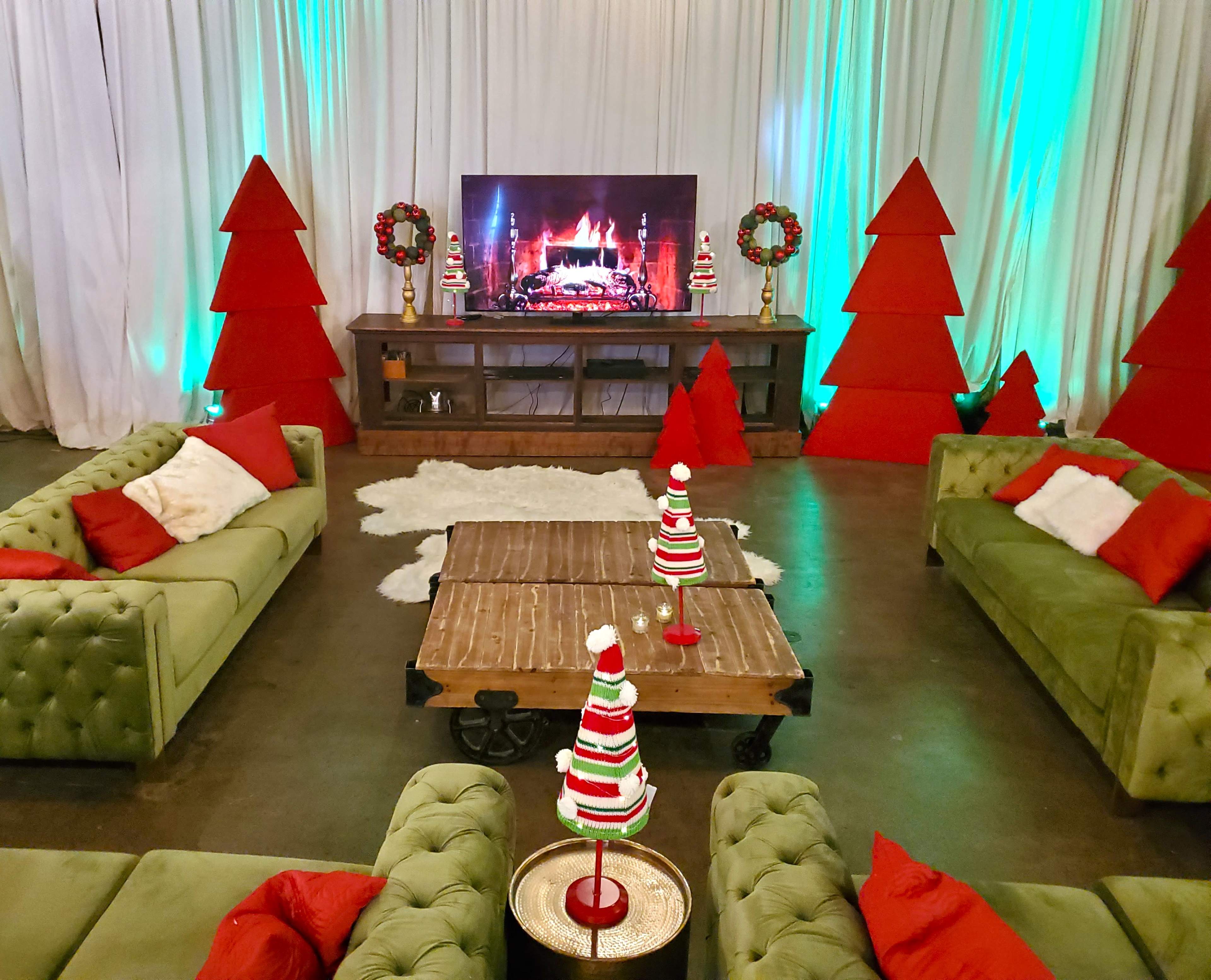 The scene features a holiday-themed room with green sofas, red and green decorative trees, and a television displaying a fireplace, all arranged around a wooden coffee table.