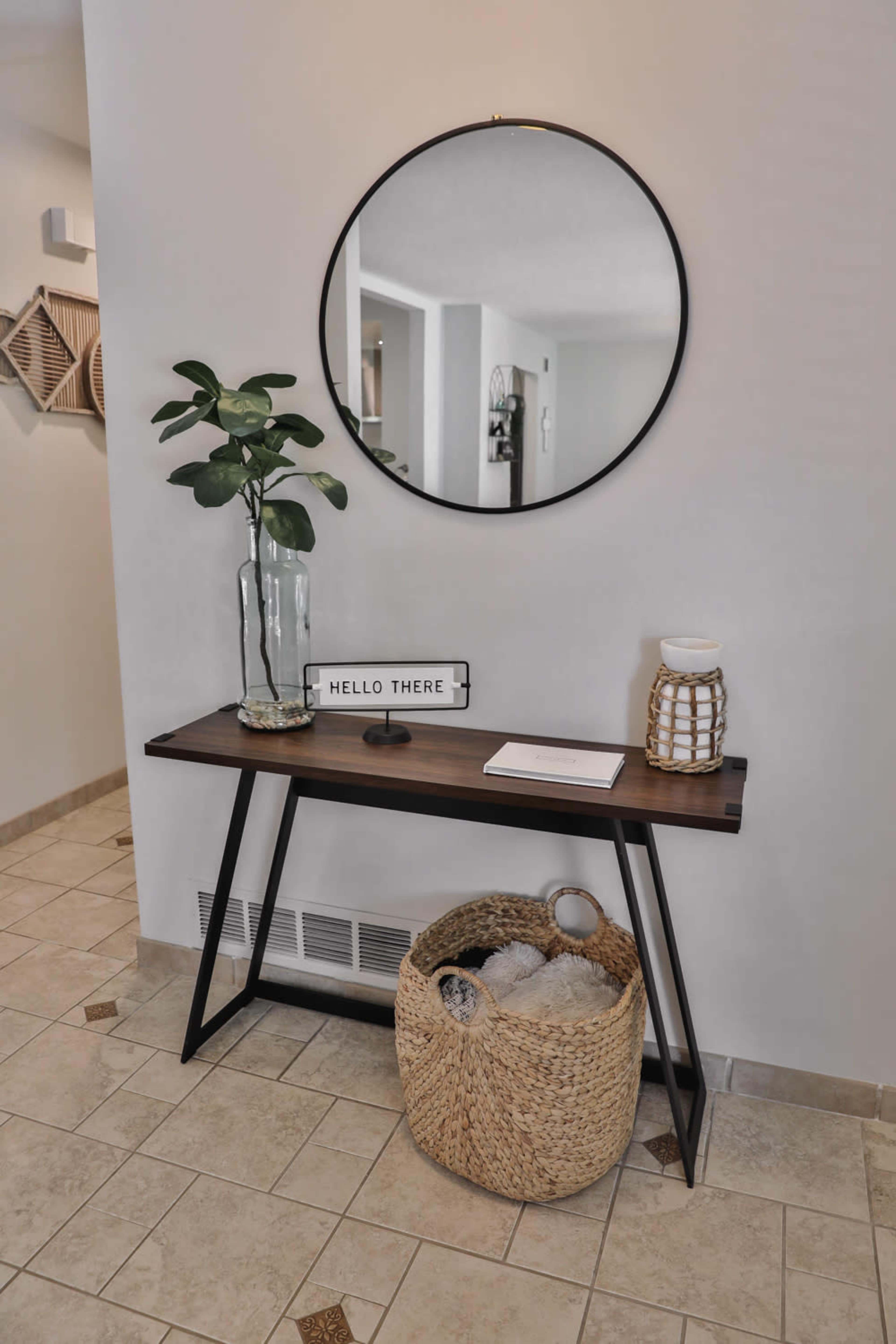A wooden console table with a round mirror above it is positioned against a light-colored wall, accompanied by a plant, decorative items, and a woven basket.